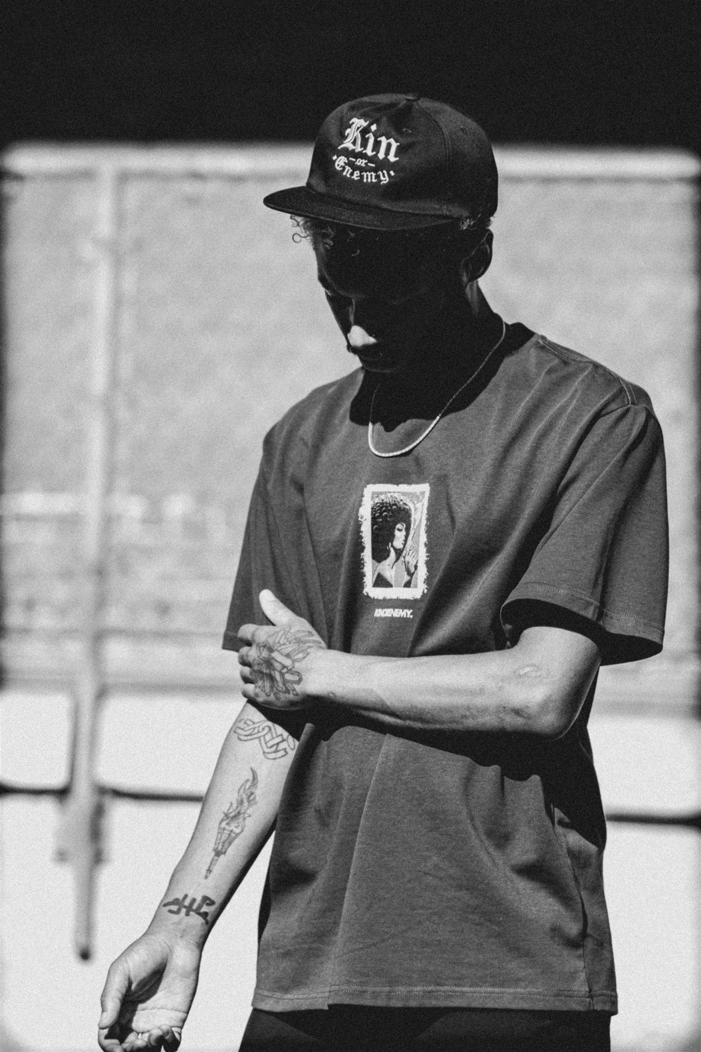 A person wearing a baseball cap with the text 'Fan or Enemy' and a graphic T-shirt, standing outdoors with their arms crossed, tattoos visible on their tattooed arm, in black and white.