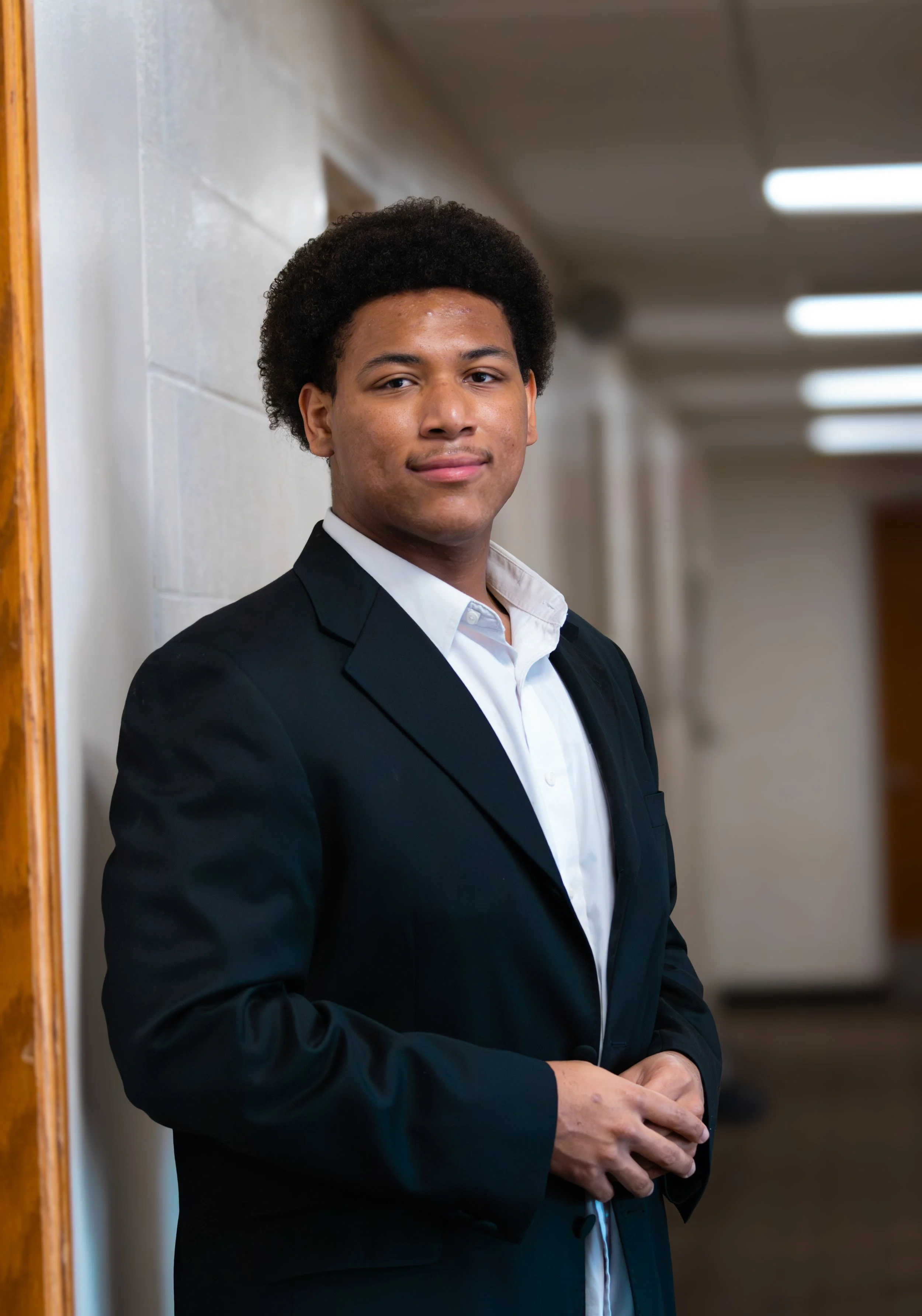 Young man in a black suit and white shirt standing in a hallway, looking at the camera with a slight smile.