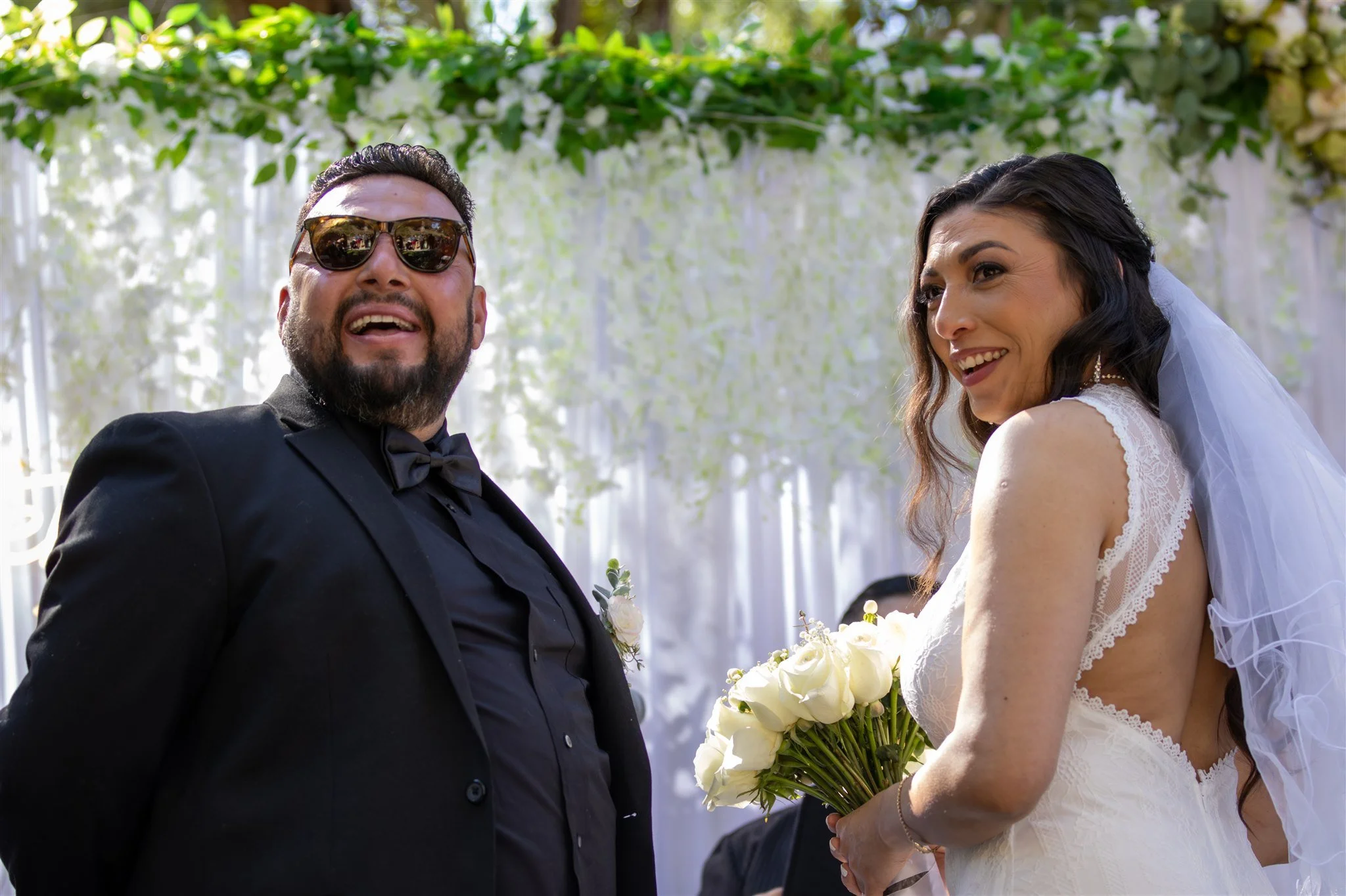 A smiling man wearing sunglasses and a tuxedo stands next to a woman in a wedding dress holding a bouquet of white roses during a wedding ceremony under a floral canopy.