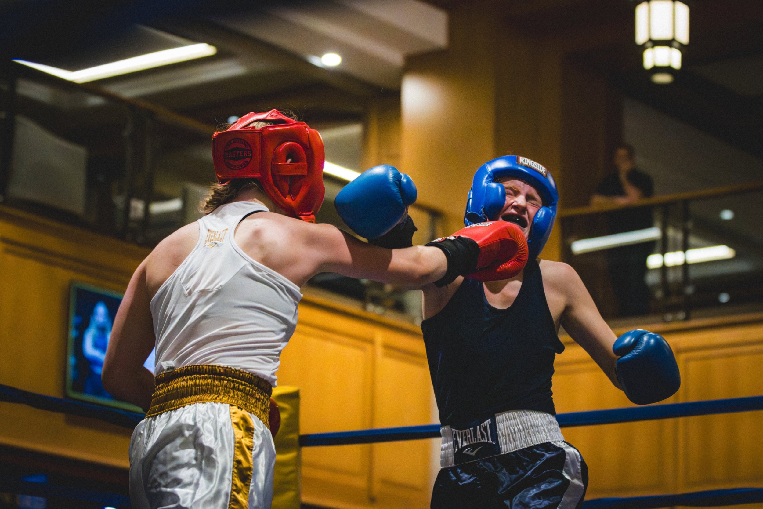 Two male boxers wearing headgear and boxing gloves sparring in a boxing ring indoors, with one throwing a punch at the other.