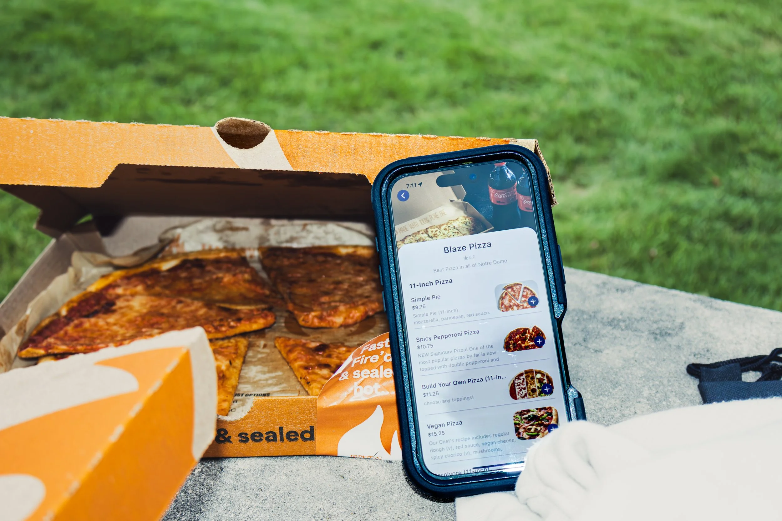 A partially opened pizza box with pizza inside, an iPhone displaying a menu for Blaze Pizza, and a grassy outdoor setting in the background.