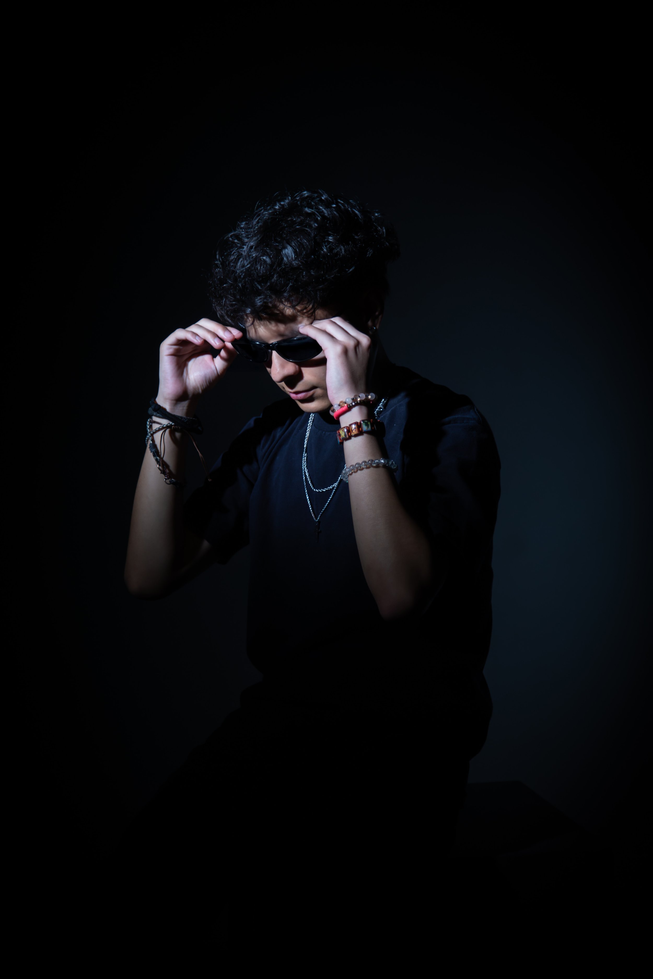 Person with dark messy hair wearing sunglasses and black clothing, adjusting sunglasses against a dark background.