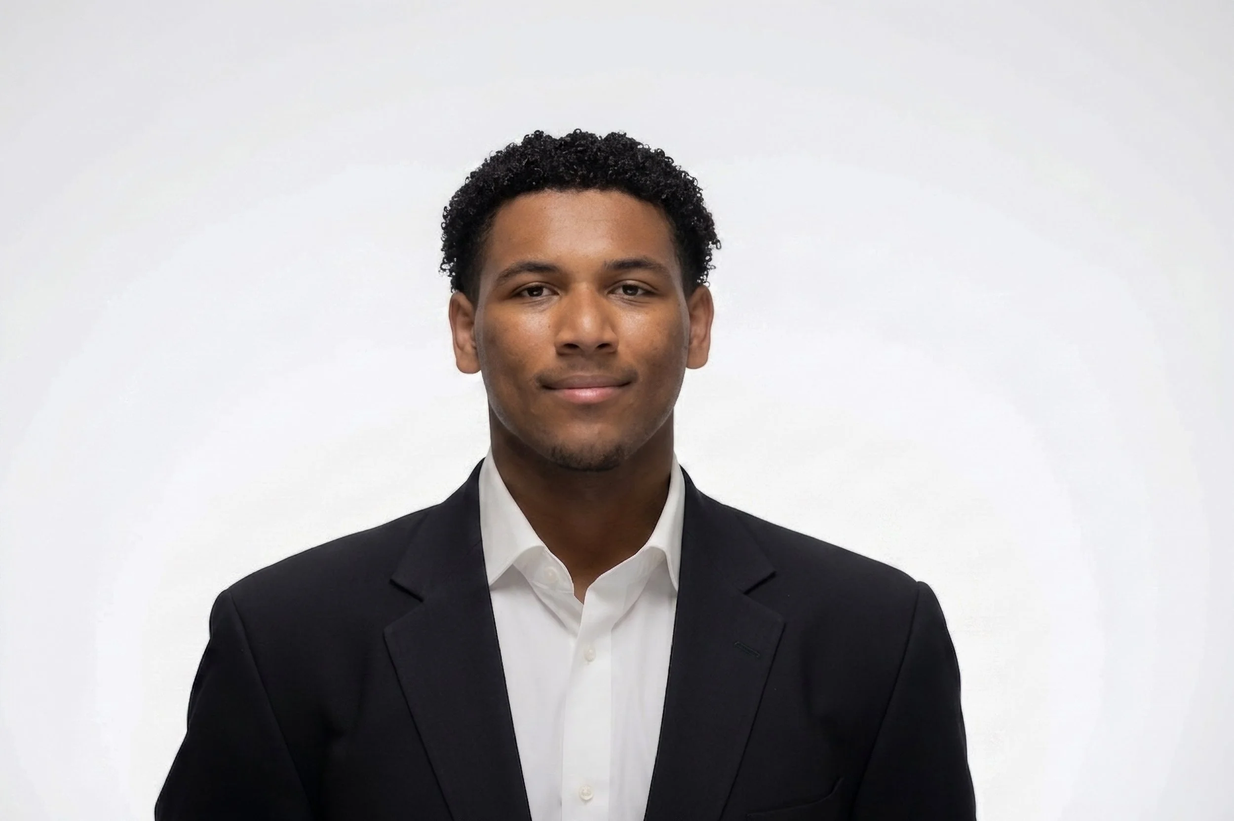 Portrait of a young man with short, curly black hair, wearing a black suit and white dress shirt, standing against a plain white background.