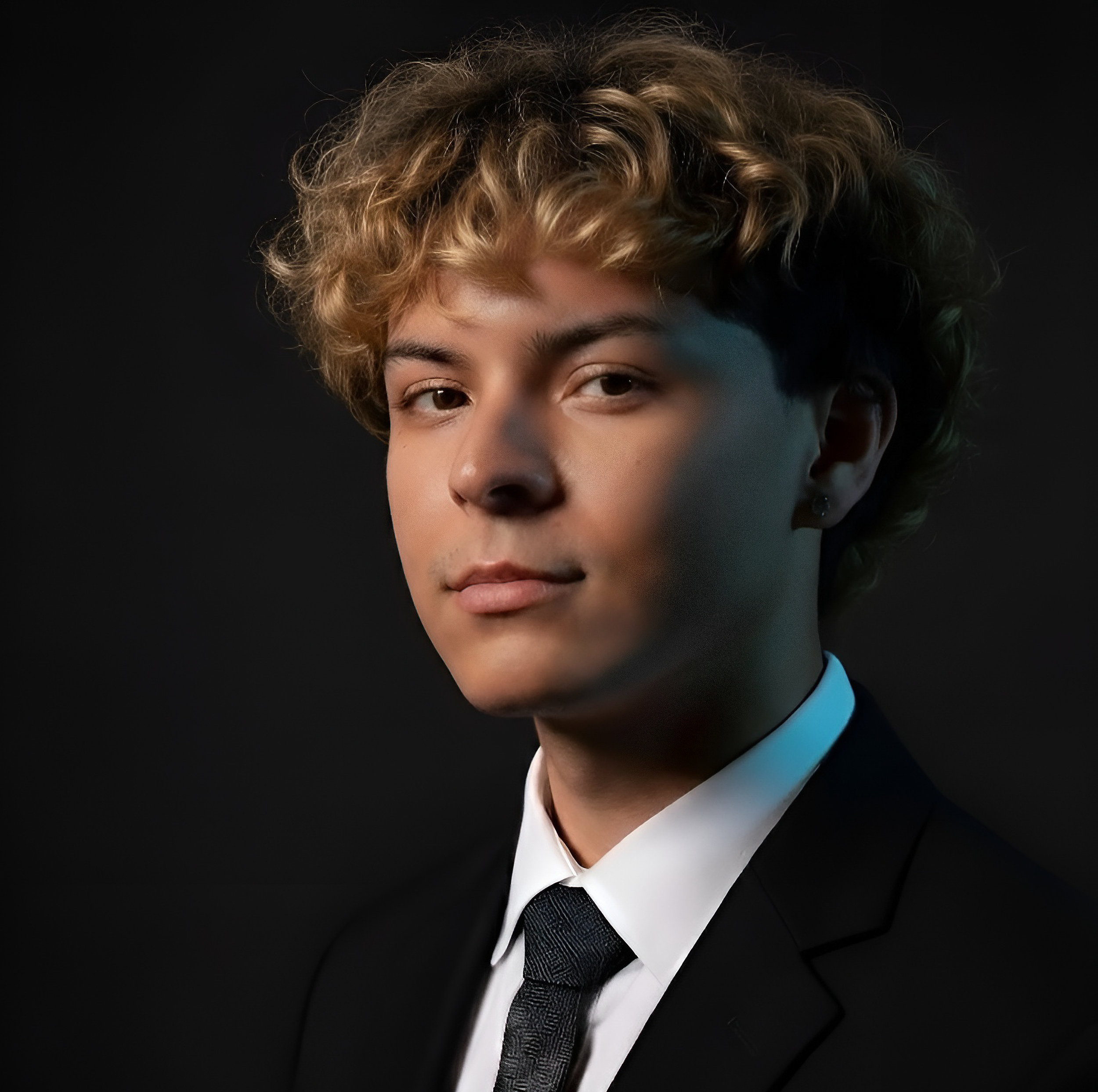 Close-up of a young person with curly hair, wearing a formal suit with a tie, against a black background.