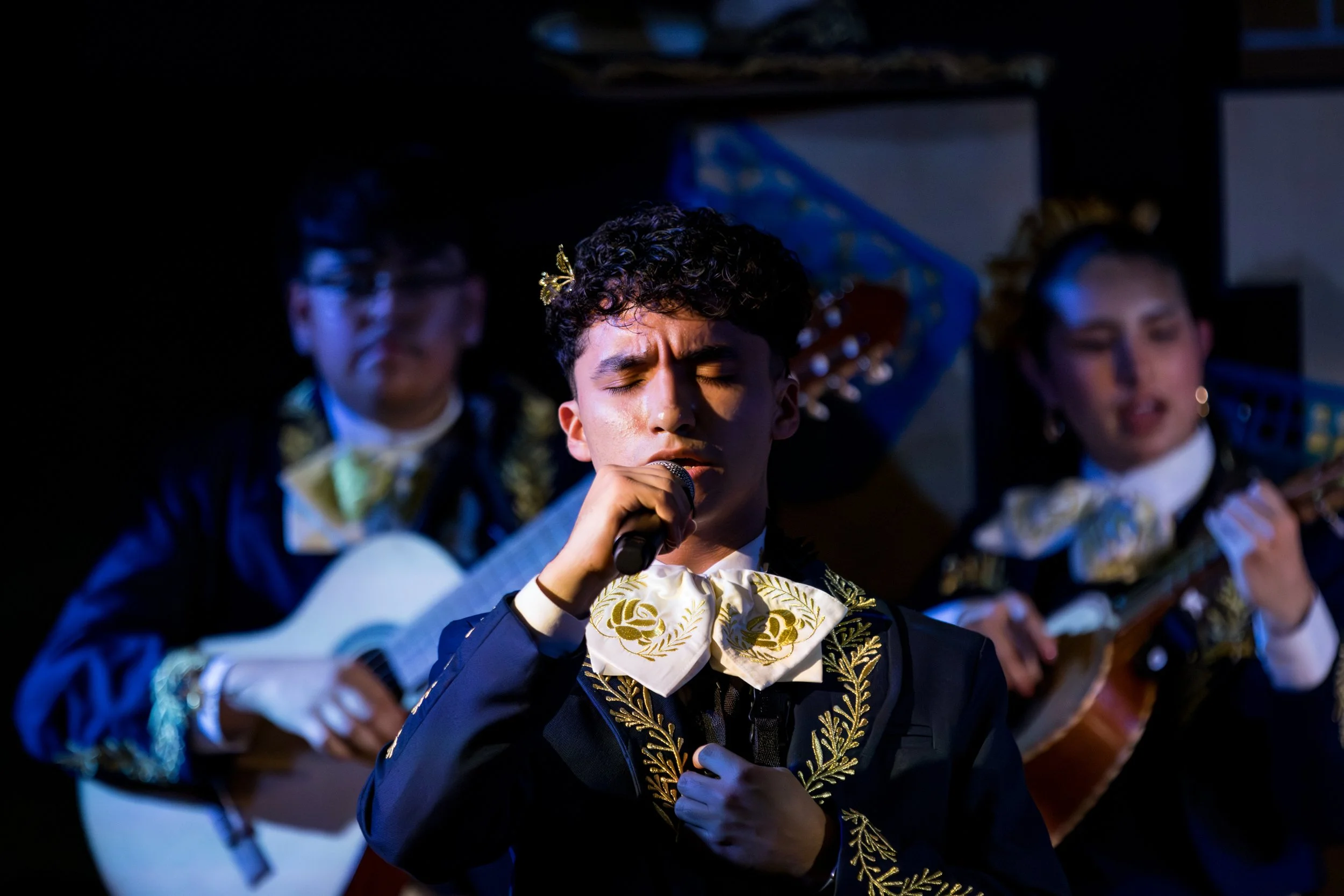 A young man with curly hair and closed eyes singing into a microphone, wearing a dark jacket with gold embroidery and a large bow tie. Two other performers are blurred in the background, one with glasses and the other with earrings, all dressed in fo