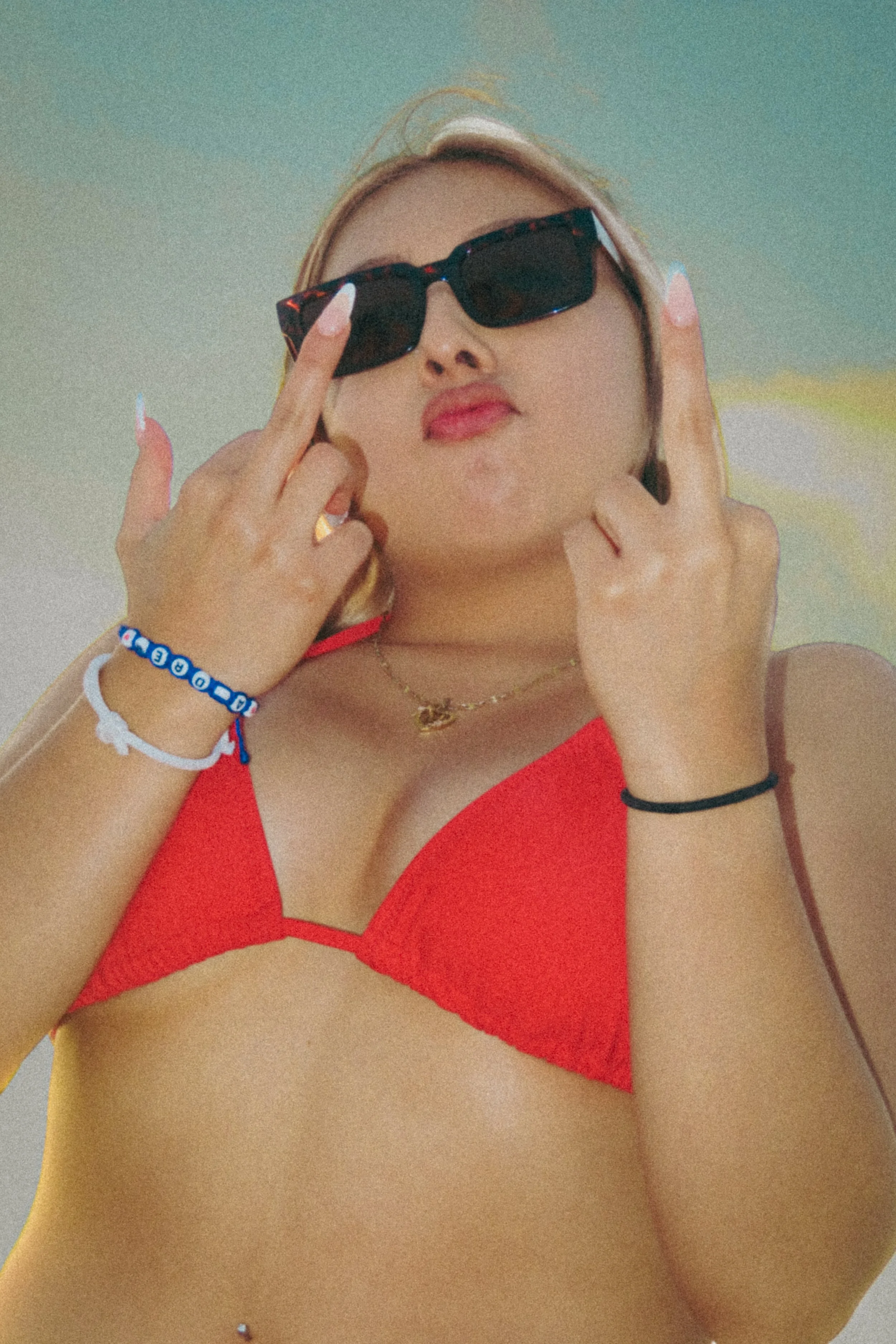A woman wearing sunglasses and a red swimsuit, pulling the sunglasses down slightly with her fingers, showing part of her face and shoulders.