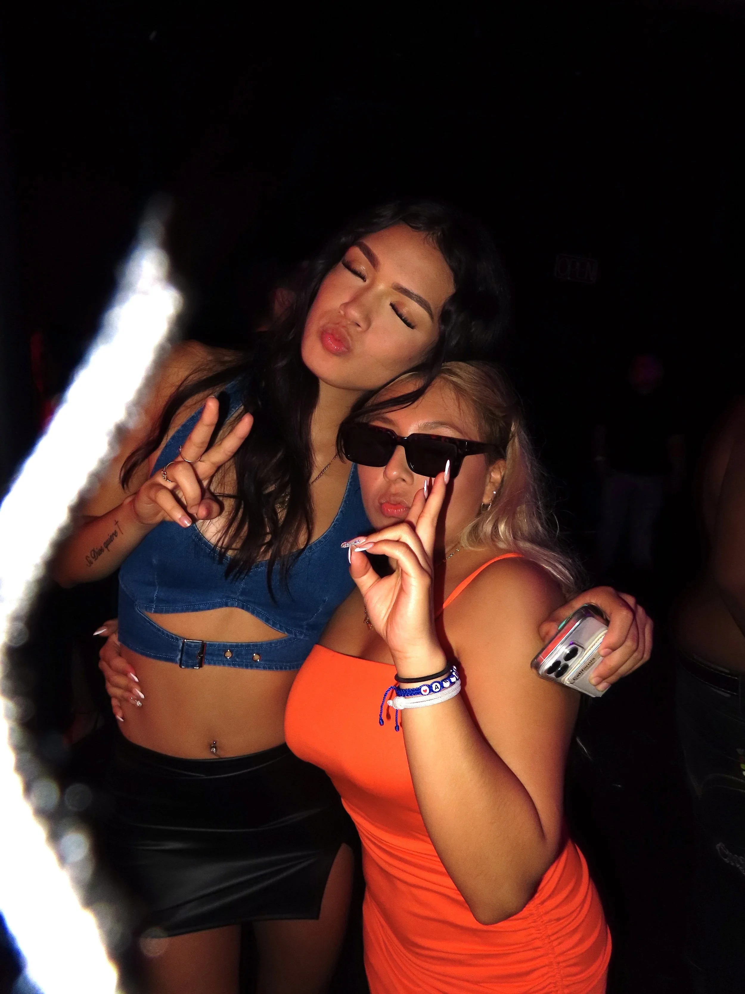 Two young women with dark and blonde hair posing together at a party, making peace signs and wearing sunglasses. One woman is wearing a blue crop top and the other an orange dress, both smiling and having fun.