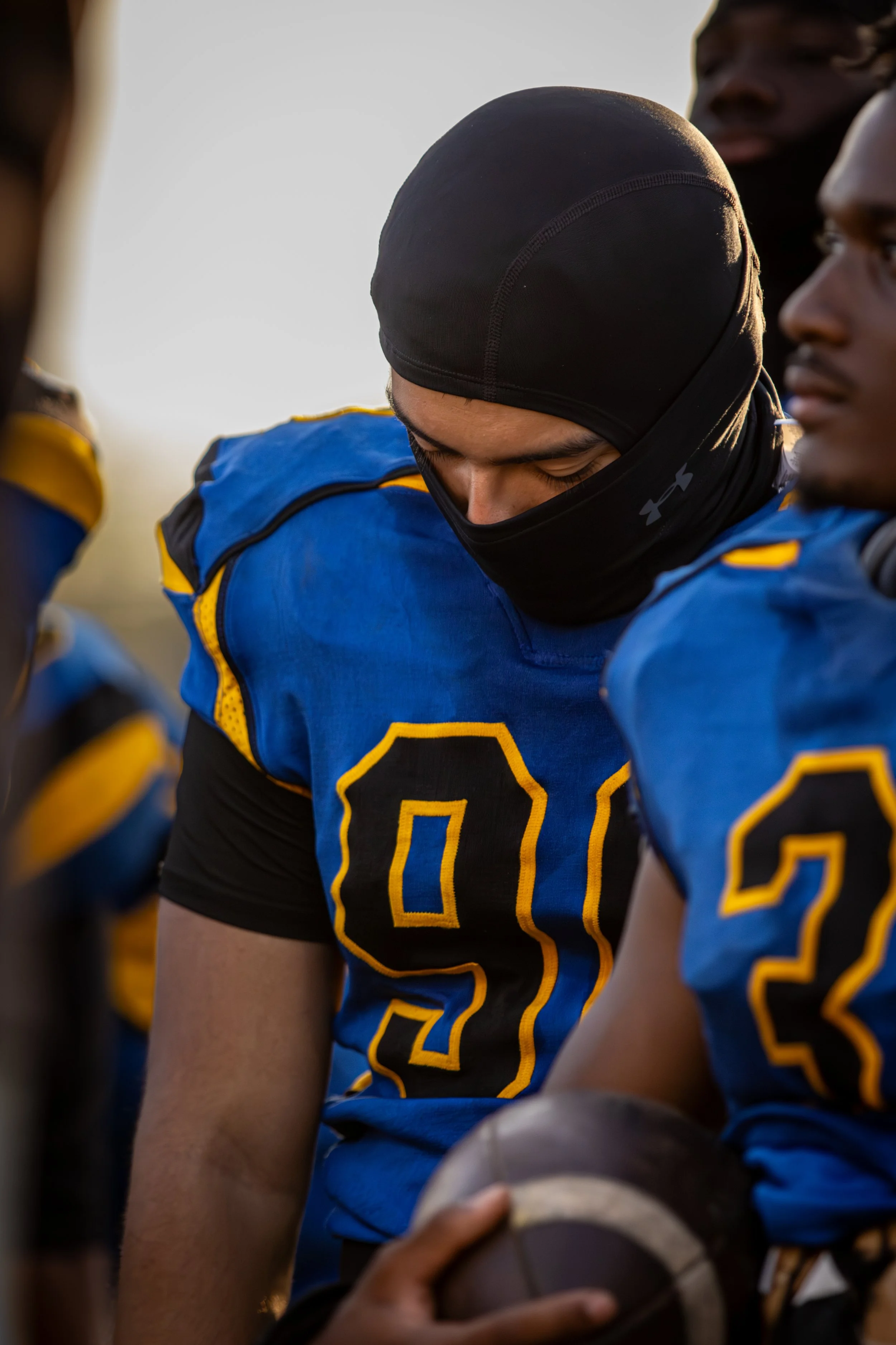 A group of football players huddled together before a game, with one player in focus wearing a blue jersey with yellow accents and the number 87, wearing a black head covering and a black face mask, holding a football.