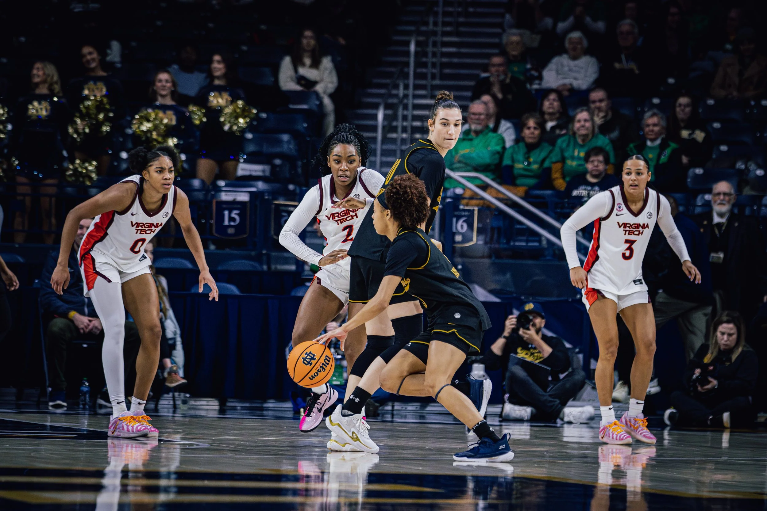 Women’s basketball game with players from Virginia Tech and an opposing team on the court, with spectators in the background.
