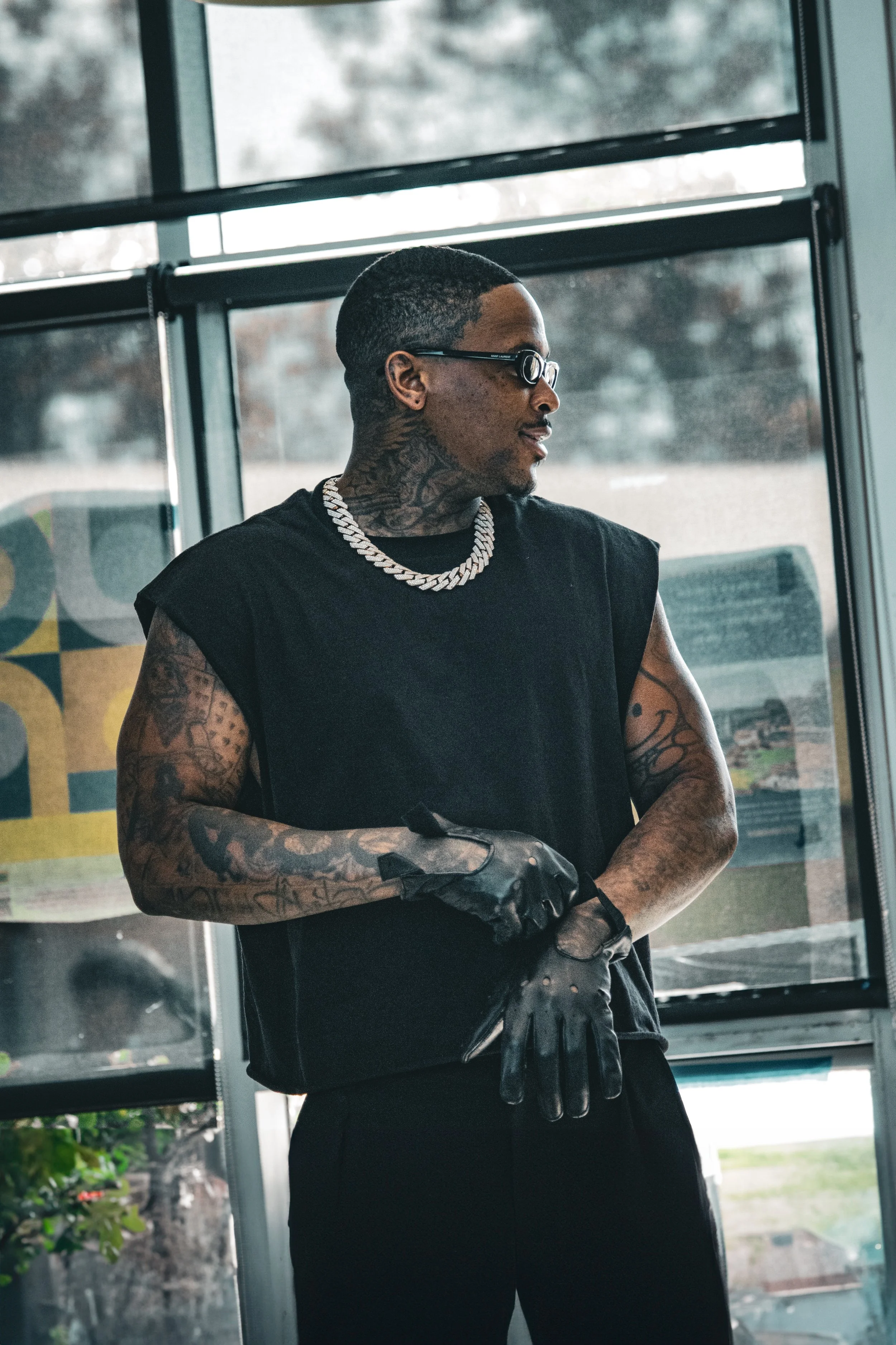 A man with tattoos on his arms and neck, wearing glasses, a black sleeveless shirt, a silver chain necklace, and black gloves, standing inside a building near a large window with a view of trees and a cloudy sky outside.