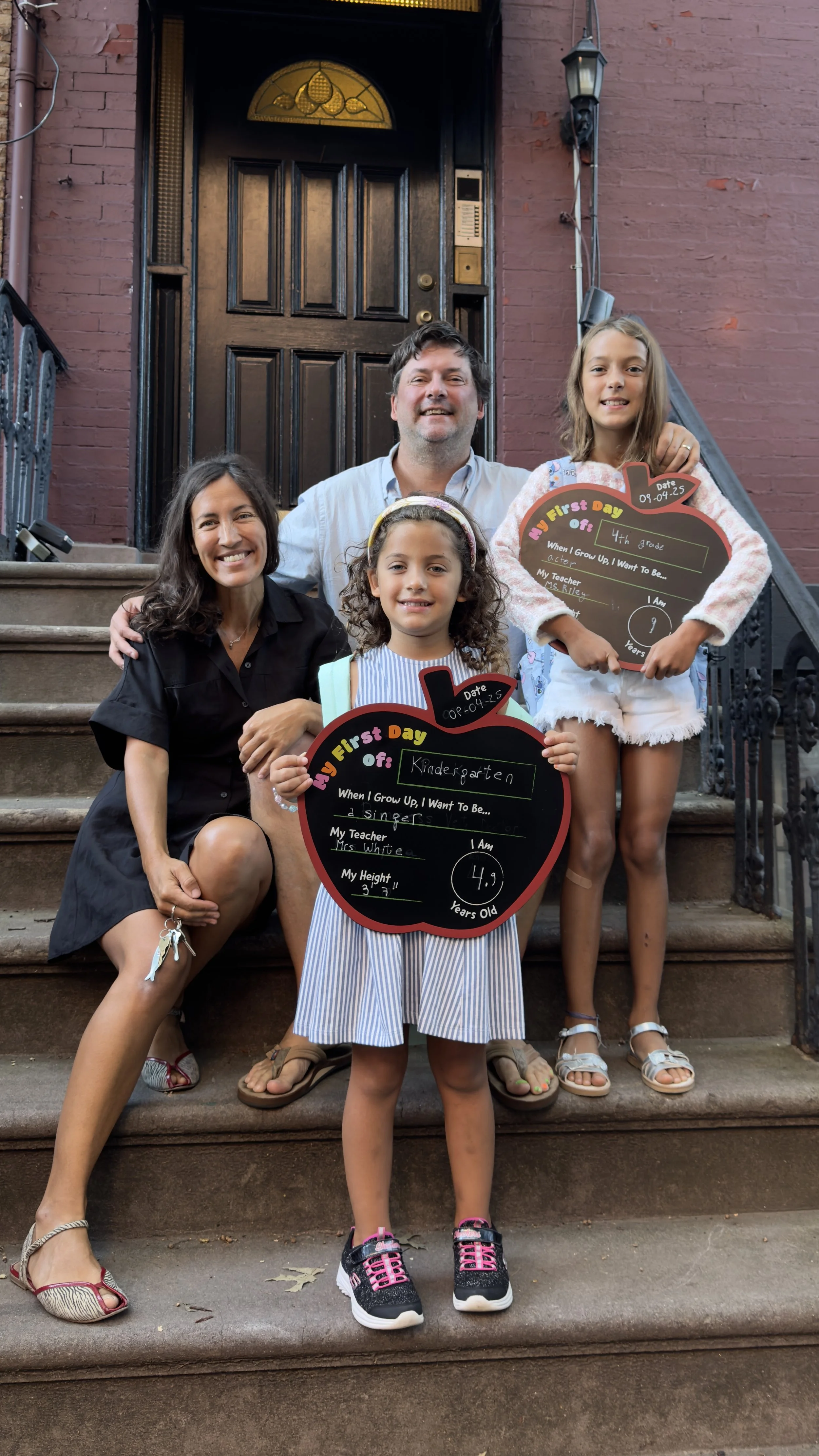 Andi Salcedo sitting with her family on a Hoboken brownstone stoop as her two daughters hold first day of school signs for Kindergarten and 4th grade.