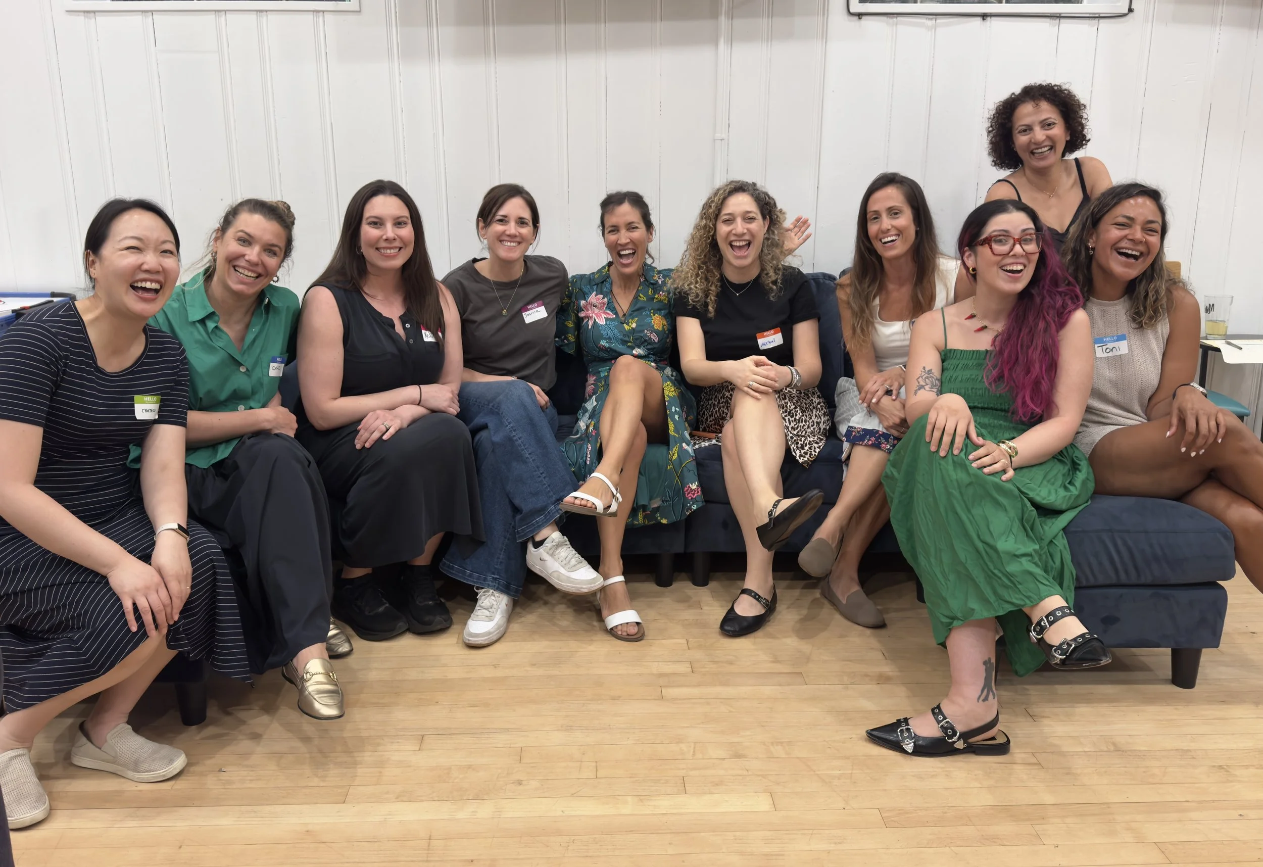 A row of women laughing genuinely while sitting close together on a couch at a local evening gathering.