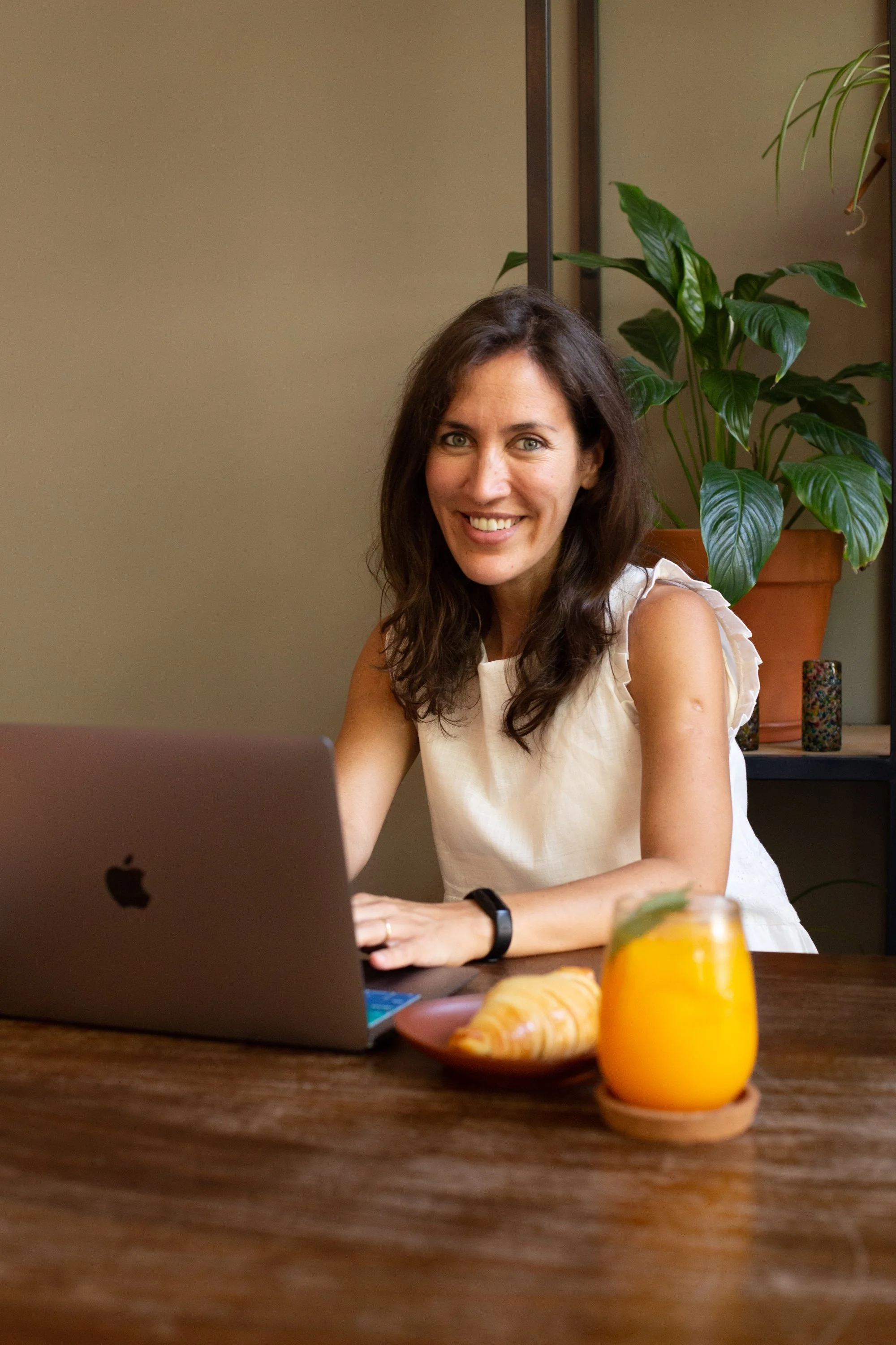 Andi Salcedo, a professional career coach for working mothers, smiling at her desk while working on a laptop in a bright office space.
