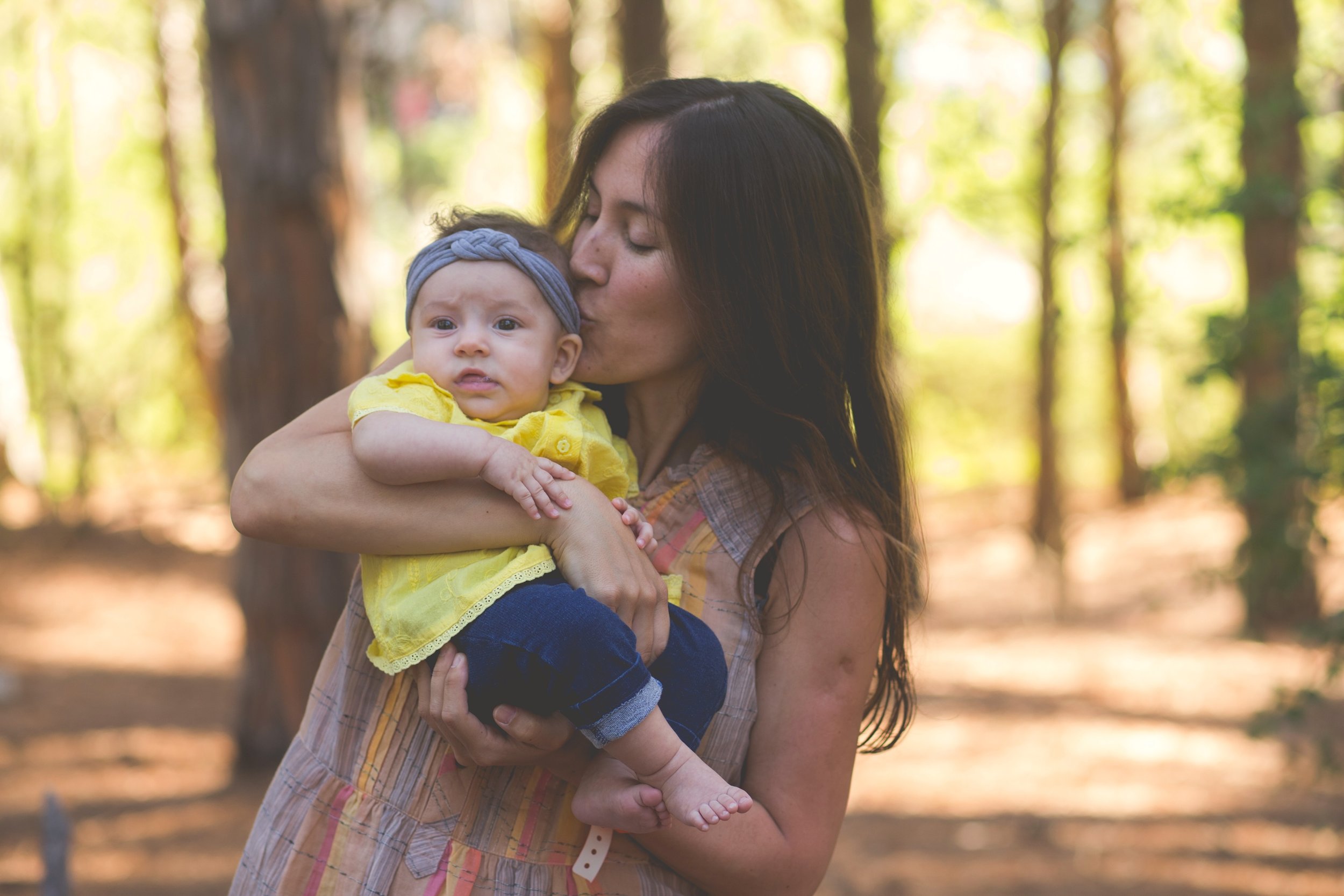 A mother kissing her baby daughter's cheek in a bright, sunlit forest setting, illustrating a sense of calm and confidence in motherhood.