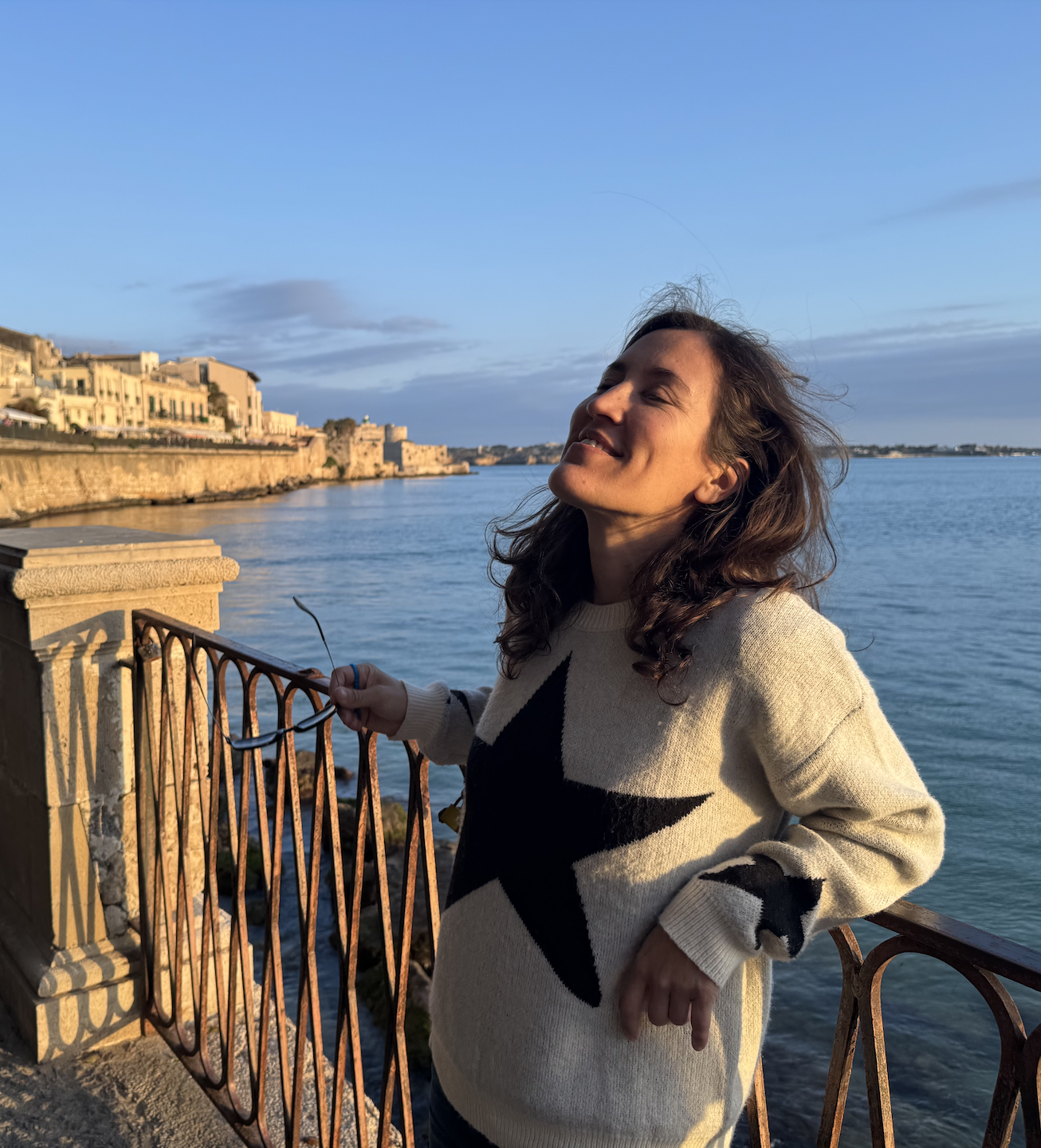 Andi Salcedo smiling with her eyes closed during a solo trip along a historic coastal stone railing, representing identity reconnection and finding clarity.