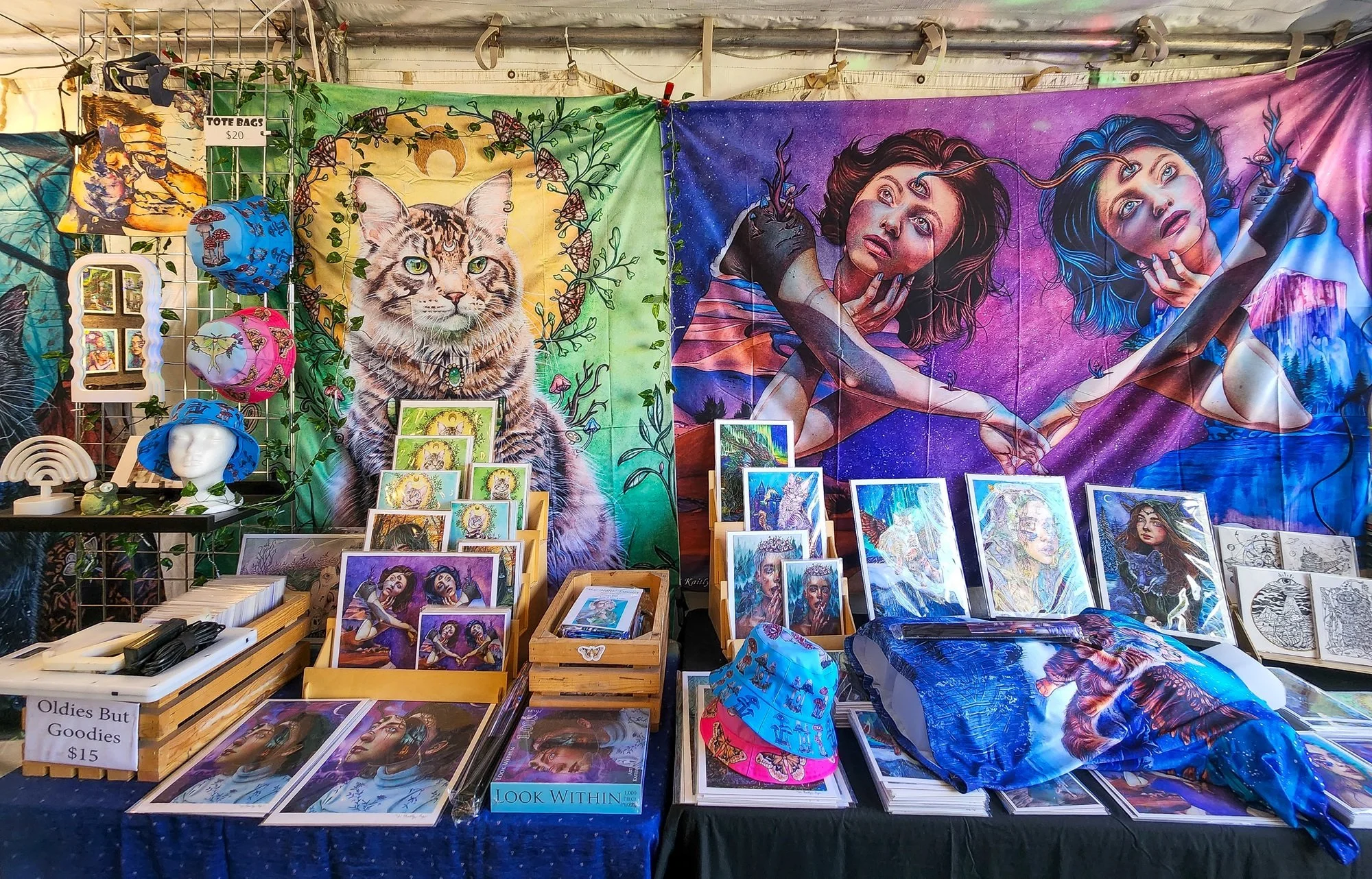Art booth displaying colorful tapestries, framed artwork, and hats. Prominent tapestry features a large tiger in front of a green and yellow background, while another depicts two women reaching out to each other against a pink and purple sky. Items are arranged on tables and a grid, with a sign reading 'Oldies But Goodies $15'.