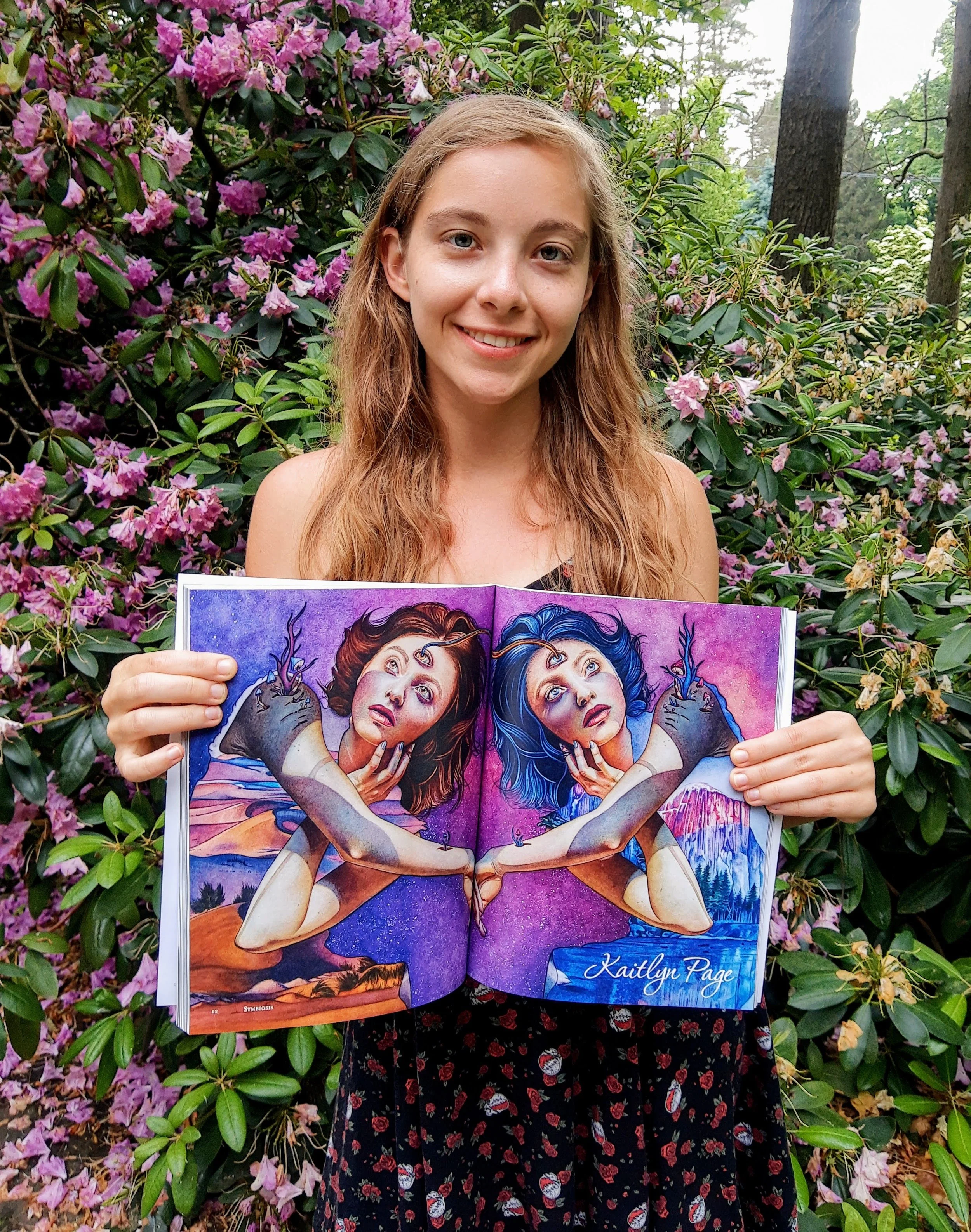 A young woman with long, wavy, light brown hair standing outdoors in front of a bush with pink flowers, holding open a colorful illustrated book with a mirrored image of two women with blue and red hair, with the name Kaitlyn Page on the right page.