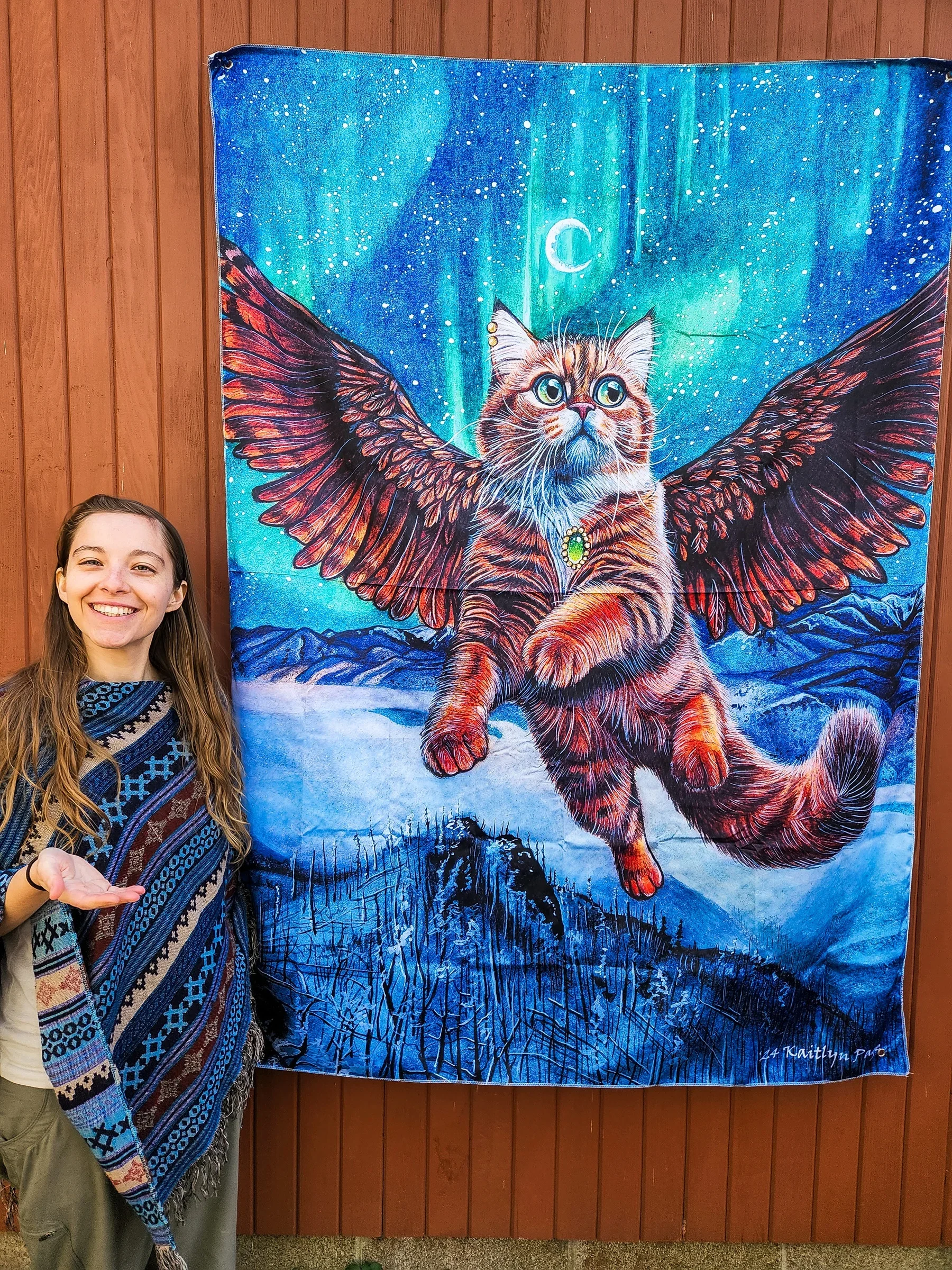 A young woman standing next to a colorful tapestry featuring a cat with owl wings, set against a night sky background with the Northern Lights and mountains.