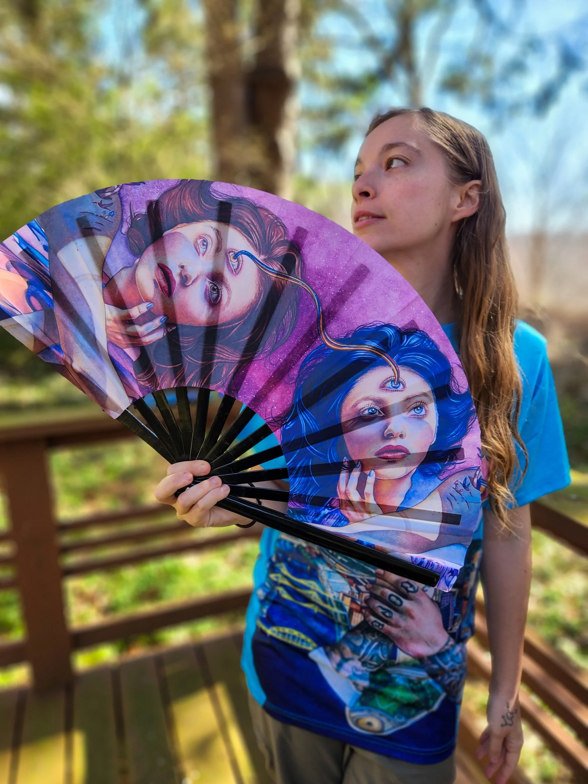 A woman with long brown hair wearing a blue shirt holds a colorful fan featuring artistic images of women with blue and purple hair, outside in a natural setting with trees and a wooden fence in the background.