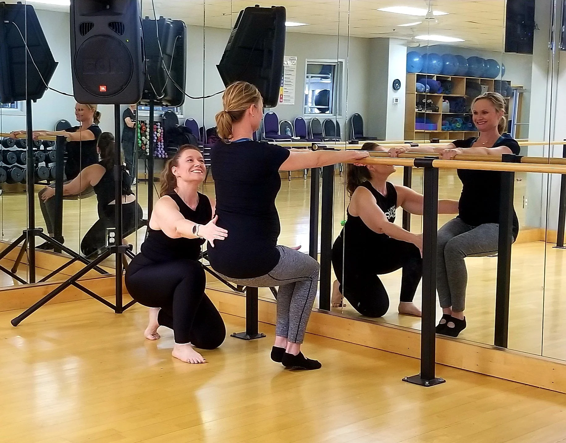 Bella and her student at the ballet barre bootybarre class with a mirror wall, wooden floors, and stacked exercise equipment in the background.