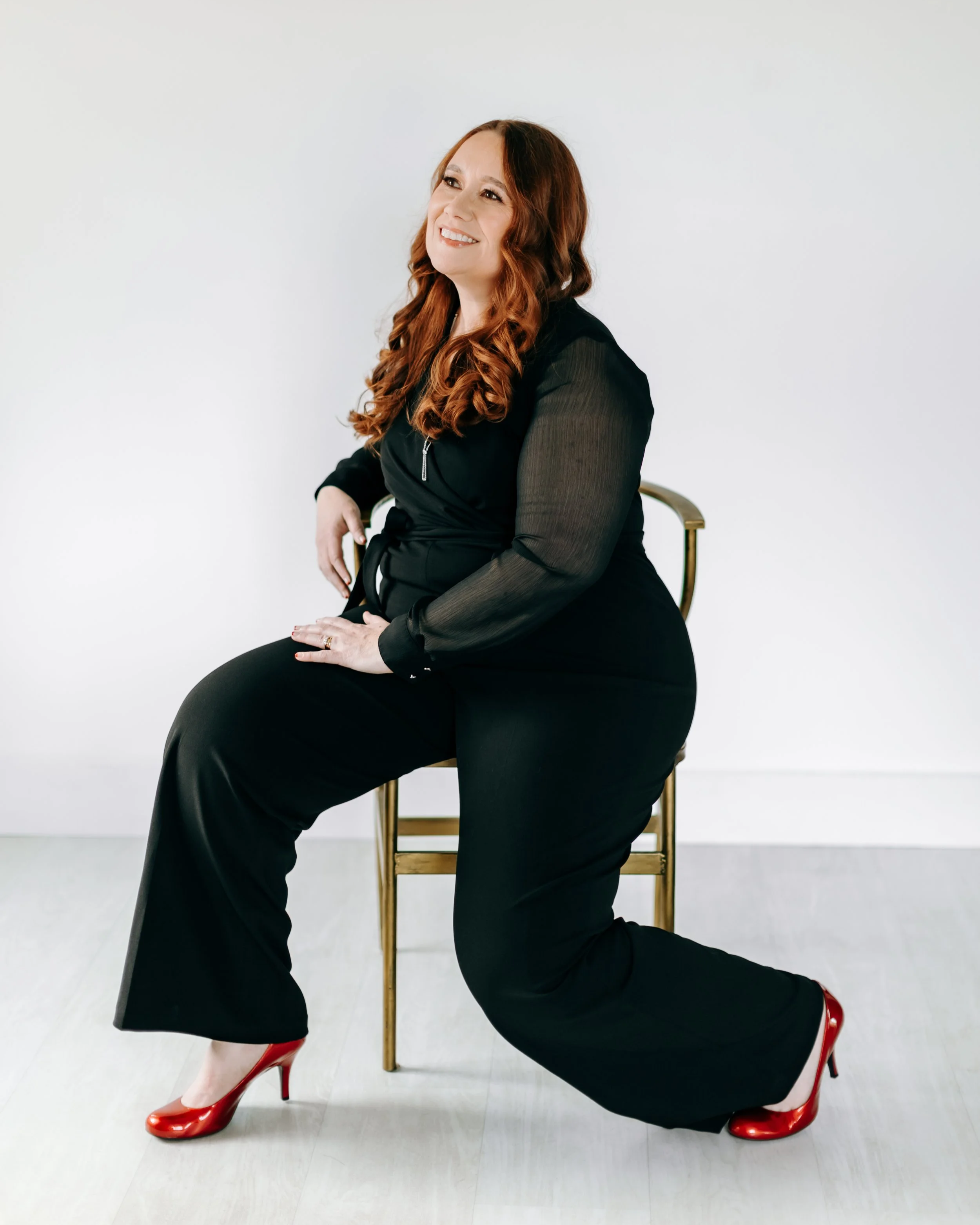 A woman in a chair looking to the side wearing a black jumpsuit and red heels.