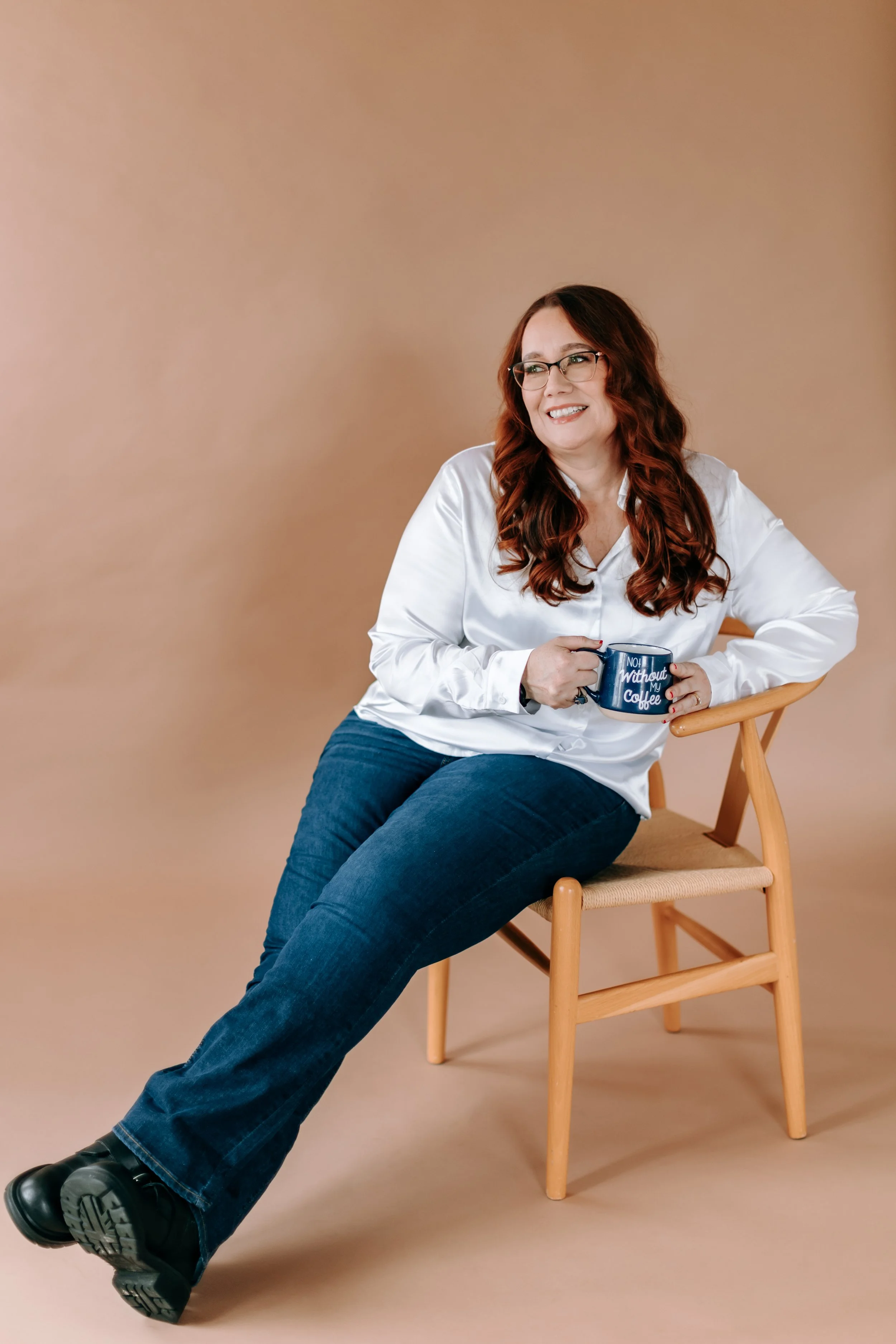 Bella. A woman with long red hair and glasses, sitting on a wooden chair against a beige background, holding a coffee mug and smiling.