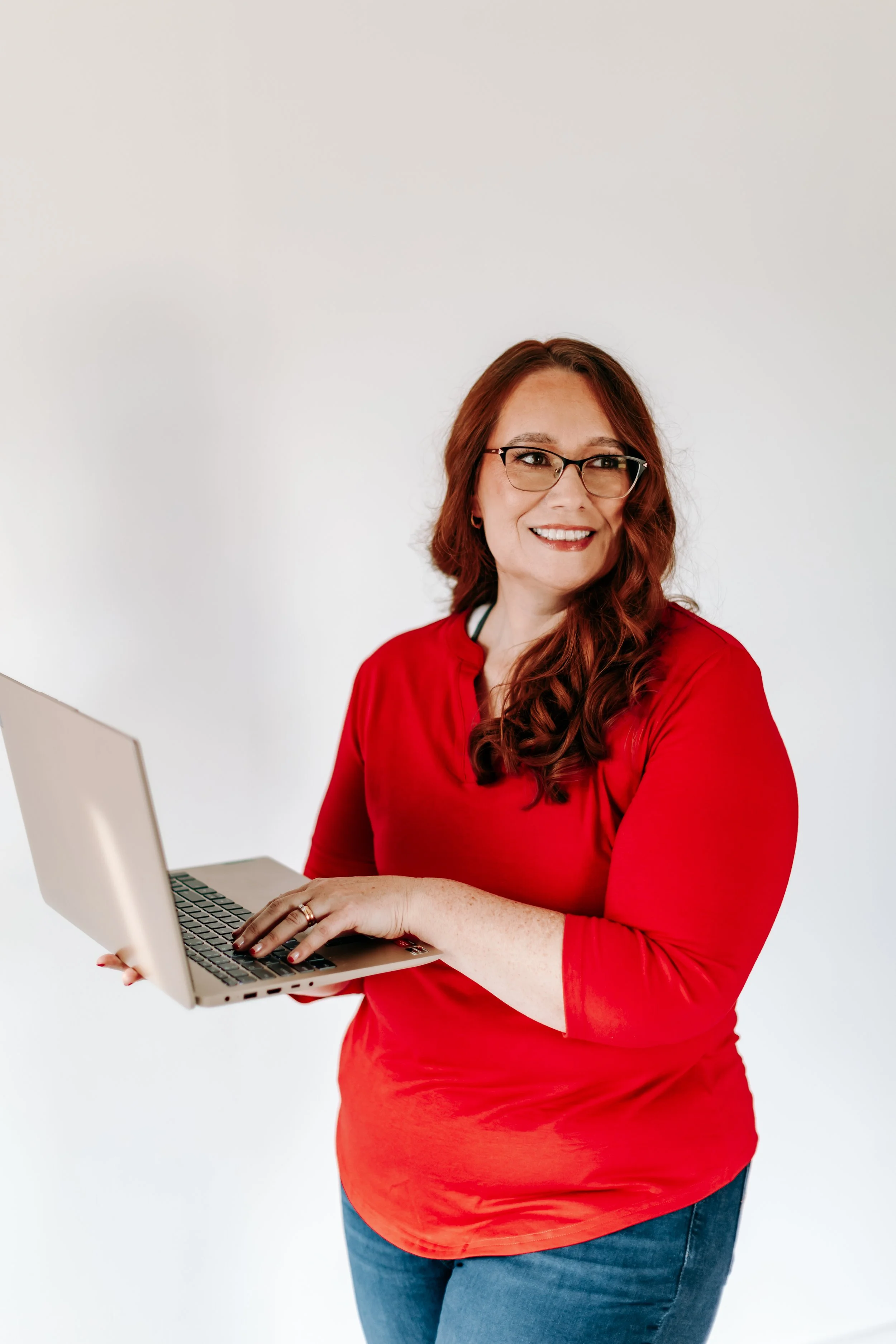 A woman with red hair, glasses, and a red shirt holding a laptop and smiling.