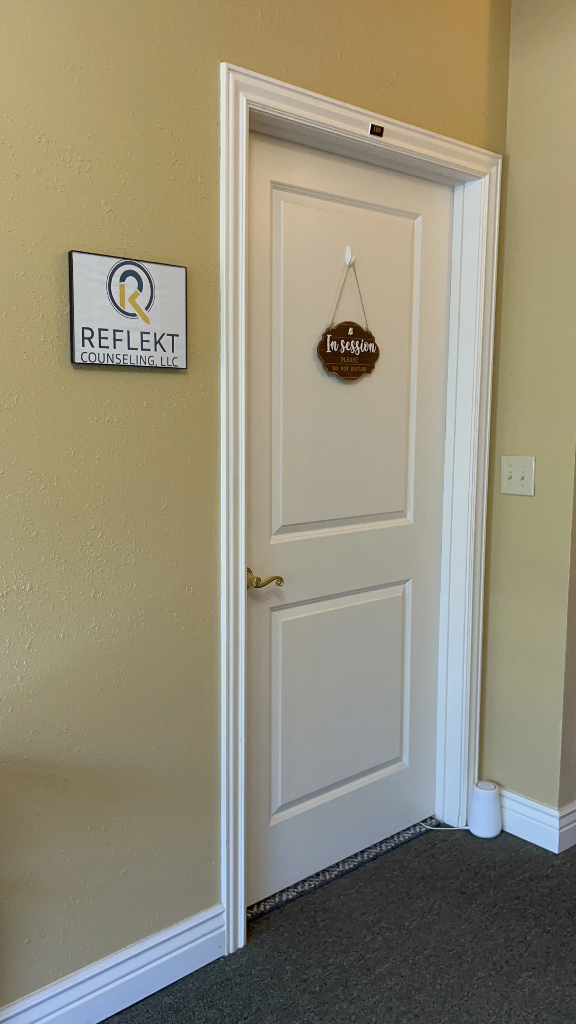 Office door with a sign that says "In session, please do not disturb," surrounded by beige walls, a sheriff's star light, and a white speaker on the floor.