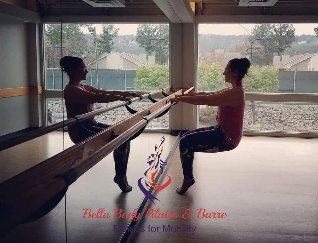A woman in a chair squat at the ballet barre facing the mirror and a open window behind her for natural light