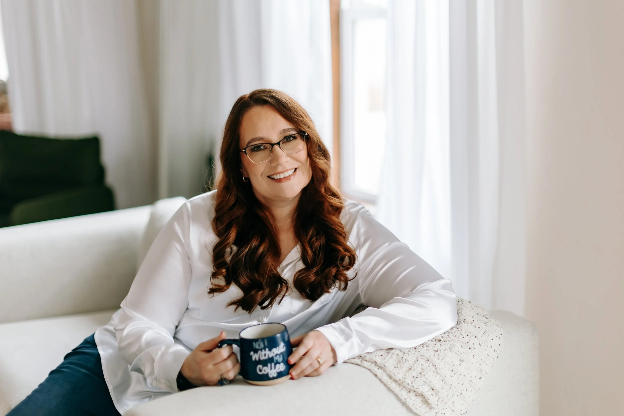 Bella, with long, wavy red hair and glasses, smiling while sitting on a white couch, holding a coffee mug that says 'Not without my coffee', in a well-lit room with sheer white curtains.