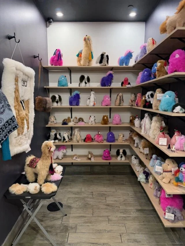 A small display room with shelves filled with colorful plush llama and alpaca stuffed animals, a few plush sheep, and accessories like hats and scarves, with a black ceiling and wood flooring.