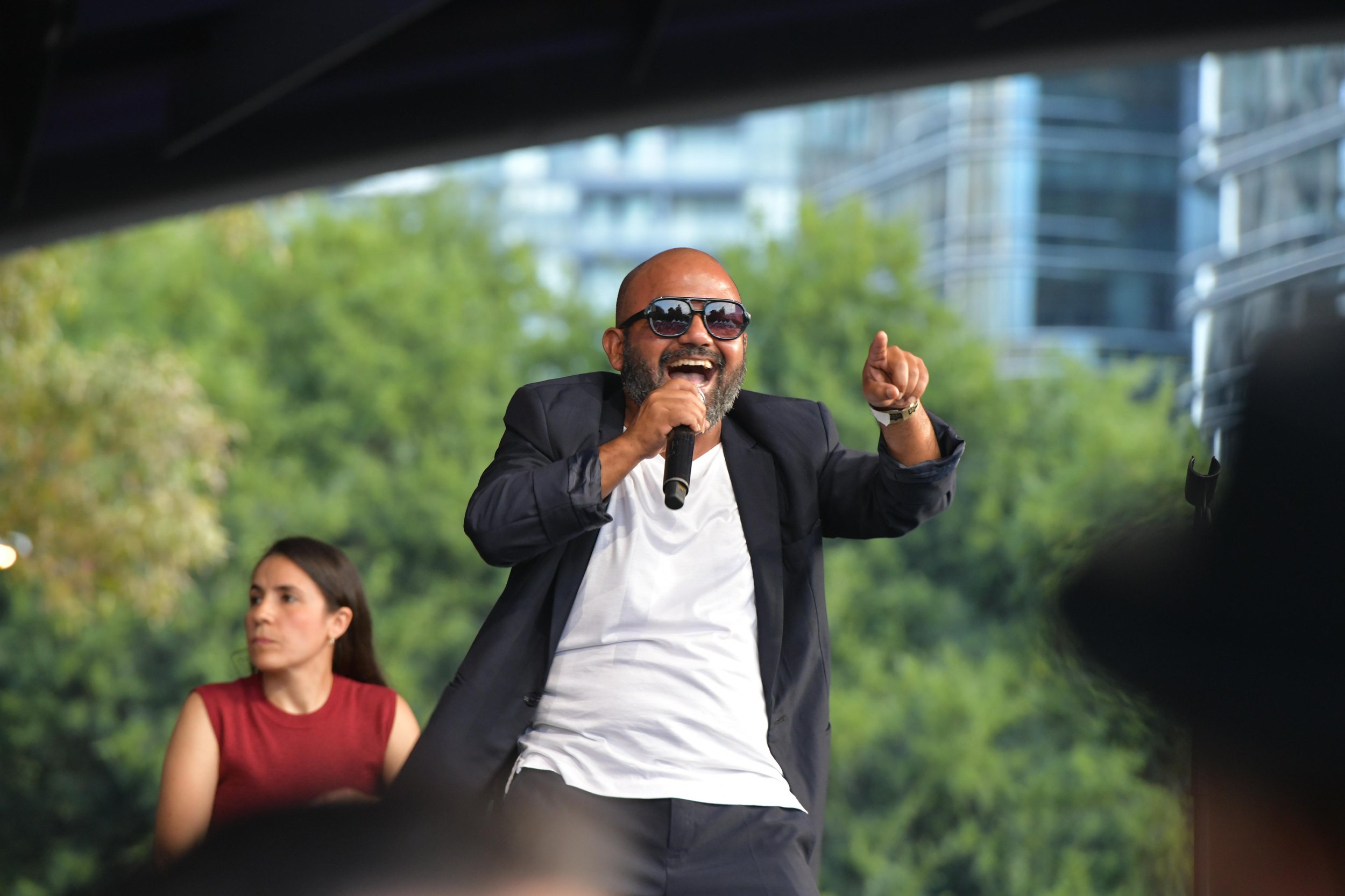 Man in sunglasses singing on outdoor stage with woman in background