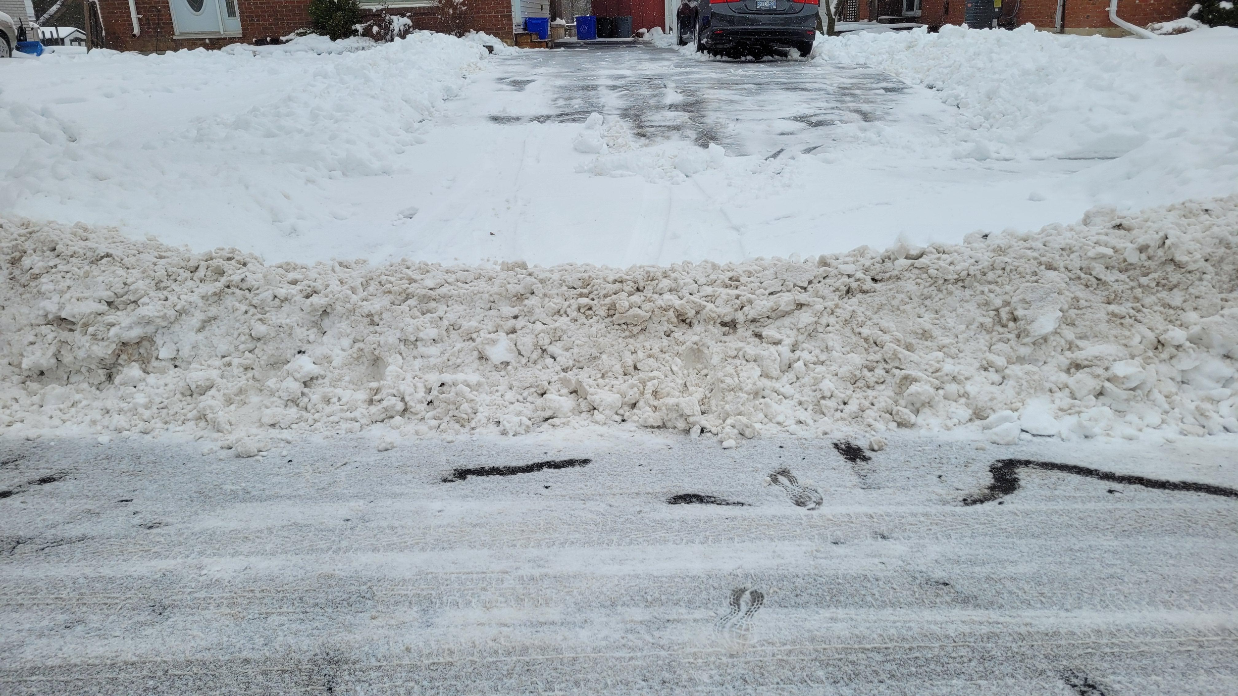 Snow piled up at the edge of a driveway with a small patch of icy pavement and footprints. In the background, a residential street with parked cars and brick houses.
