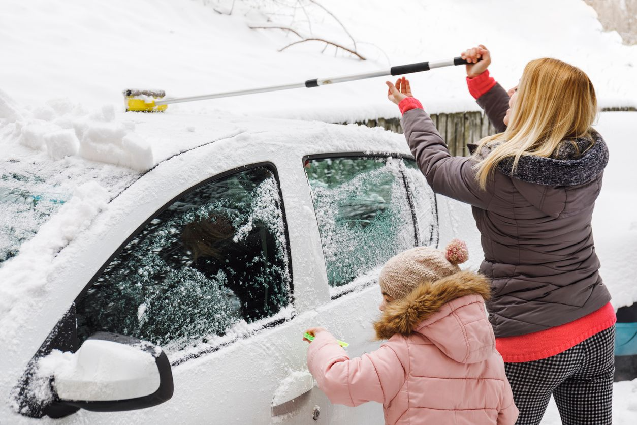 A woman and a girl cleaning snow off a white car with brushes in a snowy outdoor setting.