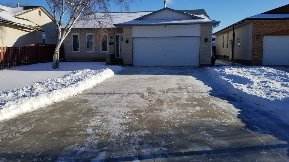 Front view of a house with a snow-covered yard and driveway, clear blue sky, and neighboring homes on each side.