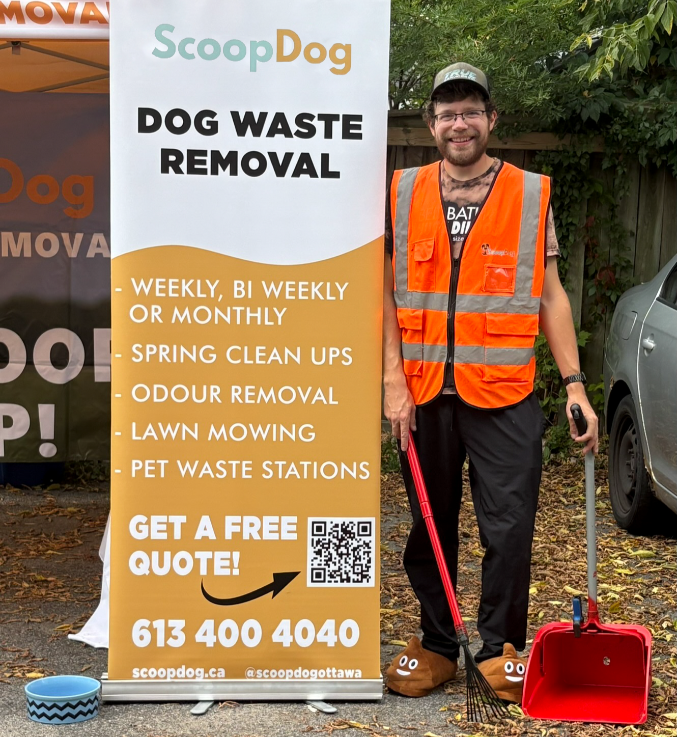 A man wearing an orange safety vest and slippers with poop emoji stands next to a large advertising banner for ScoopDog, a dog waste removal service. The banner lists services and contact information, including a QR code and a phone number, with a QR code visible in front of the man. The outdoor setting includes fallen leaves and a parked car.