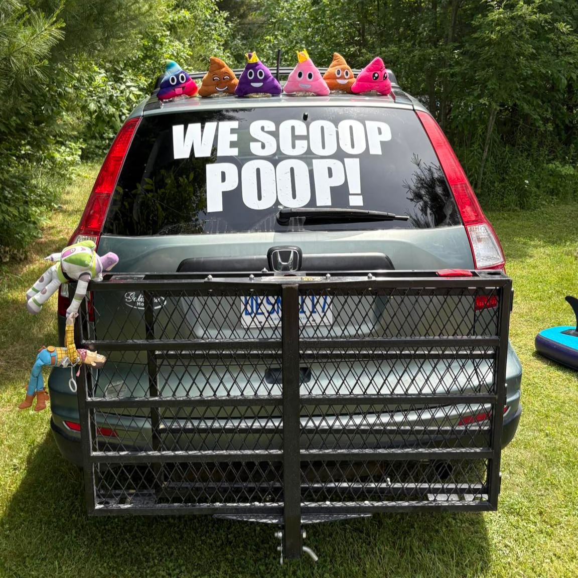 A gray Honda CR-V parked outdoors with a large black metal gate attached to the front. Plush toy characters from Toy Story, including Buzz Lightyear and Woody, are attached to the left side of the gate. Six poop emoji plush toys with crowns are placed on the top of the vehicle's rear window, and a large decal on the windshield reads "WE SCOOP POOP!" in bold white letters. The background features green trees and grass.
