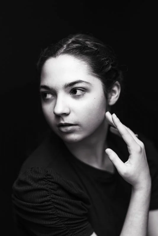 Black and white portrait of a young woman looking to the side with a thoughtful expression, hand raised near her face.