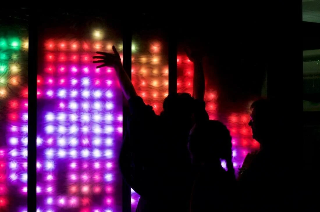 Silhouettes of people against a vibrant multicolored LED light display featuring reds, purples, and greens.