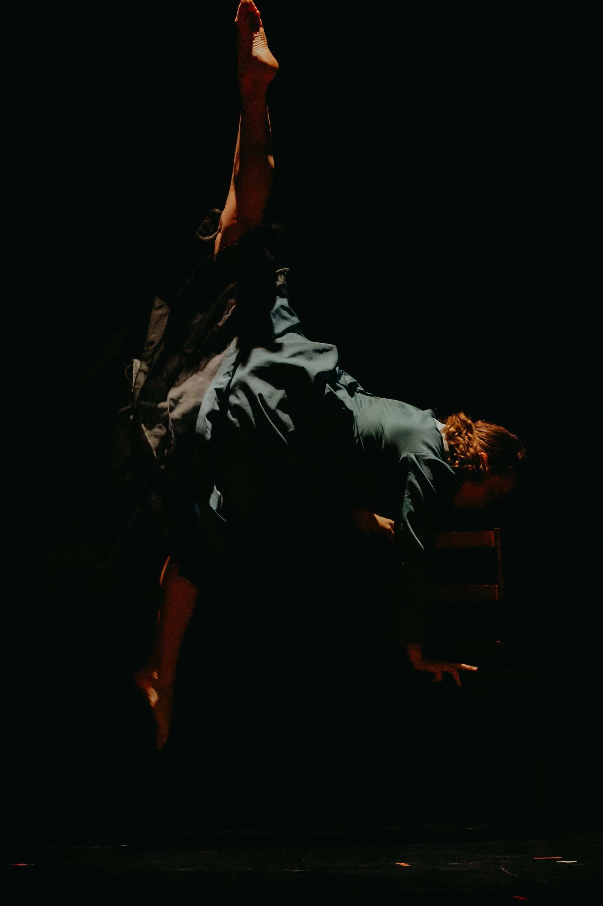 A person in a green dress performing an acrobatic dance move on a dark stage, balancing on one hand with legs extended above.