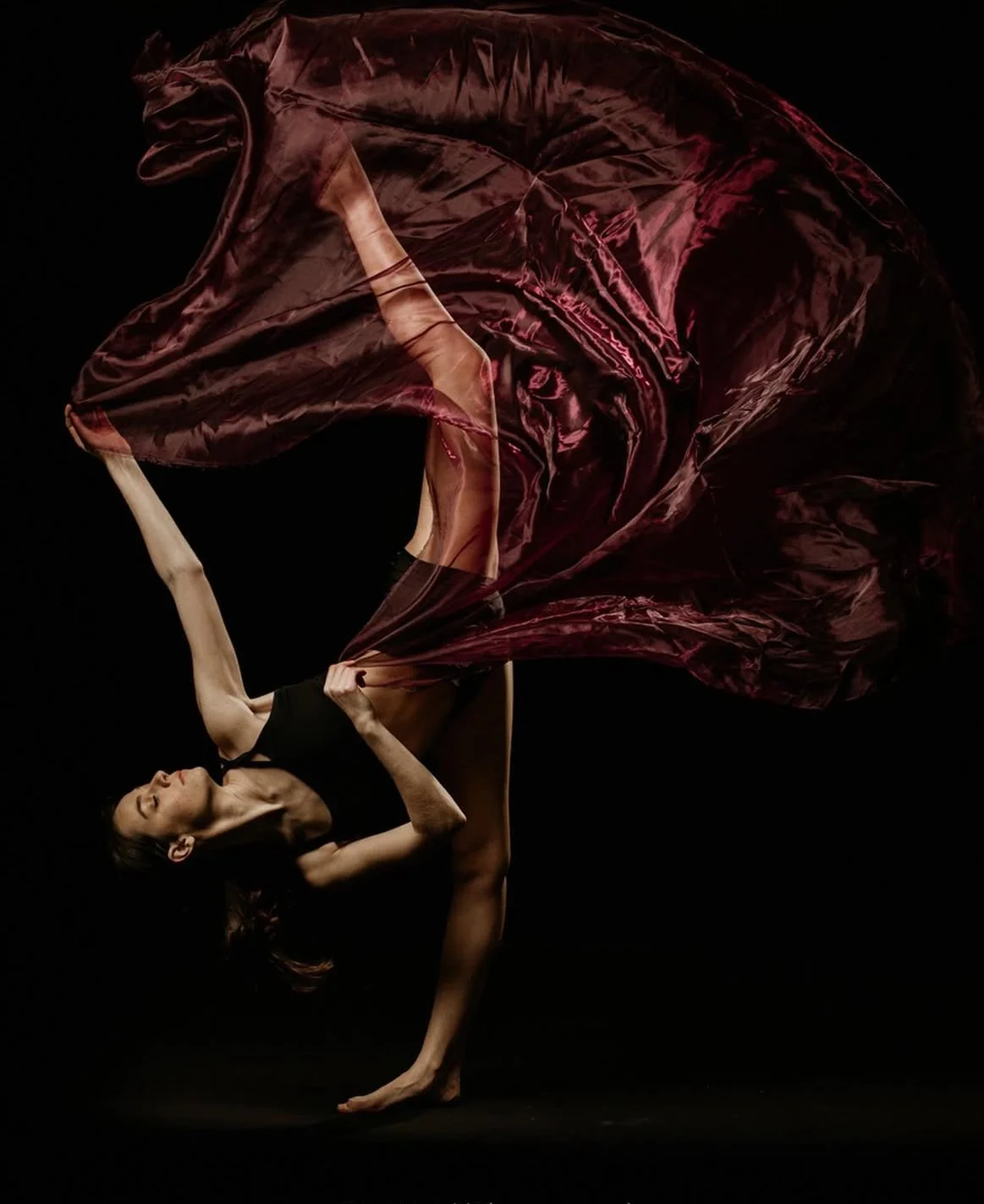 Dancer performing with a flowing maroon fabric in a dramatic pose against a dark background.