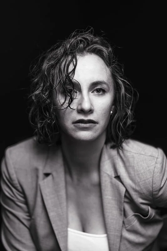 Black and white portrait of a woman with curly hair wearing a blazer, looking directly at the camera against a dark background.