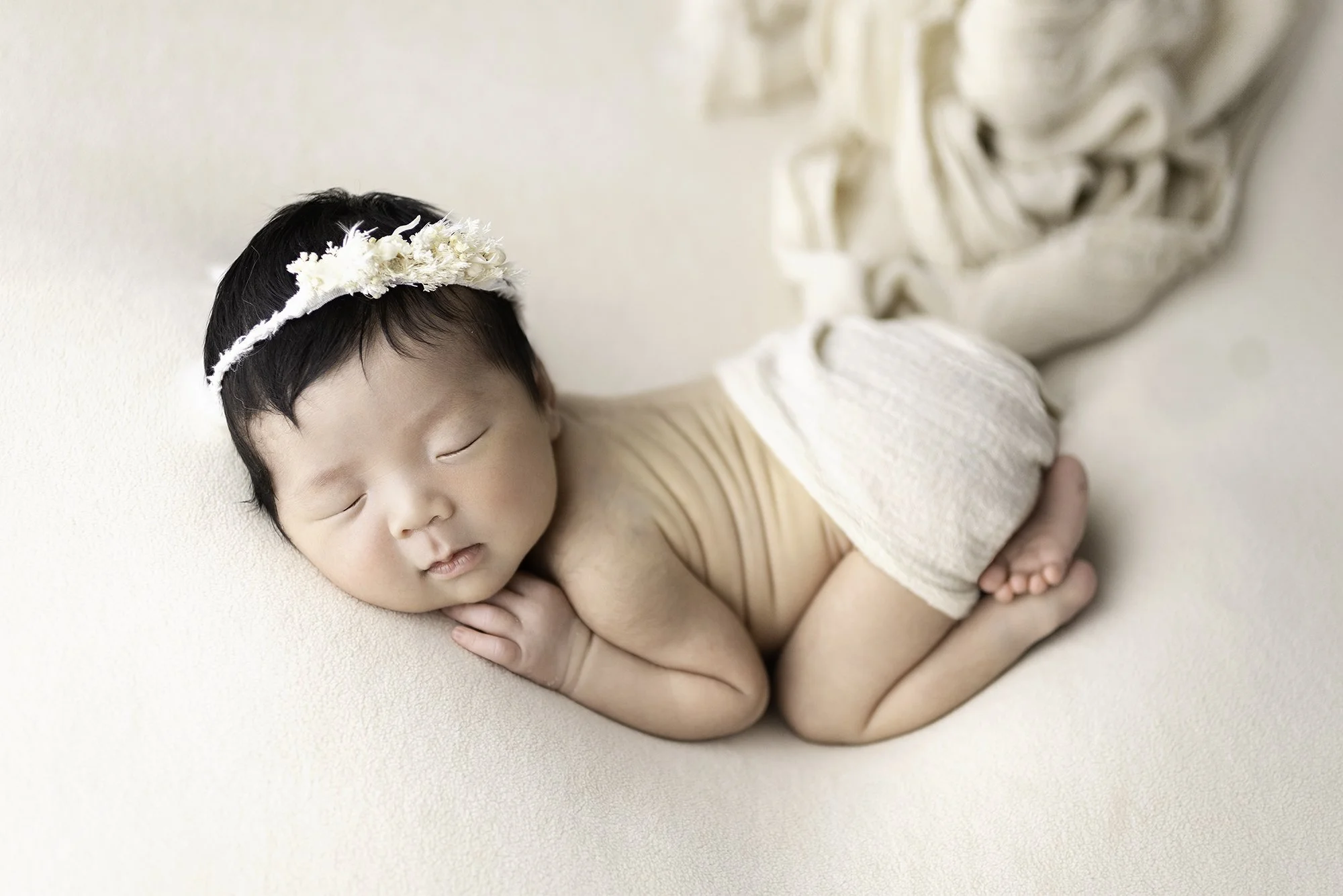 白い背景の上に寝ている赤ちゃん。花のヘアバンドをつけて、白いパンツをはいている。