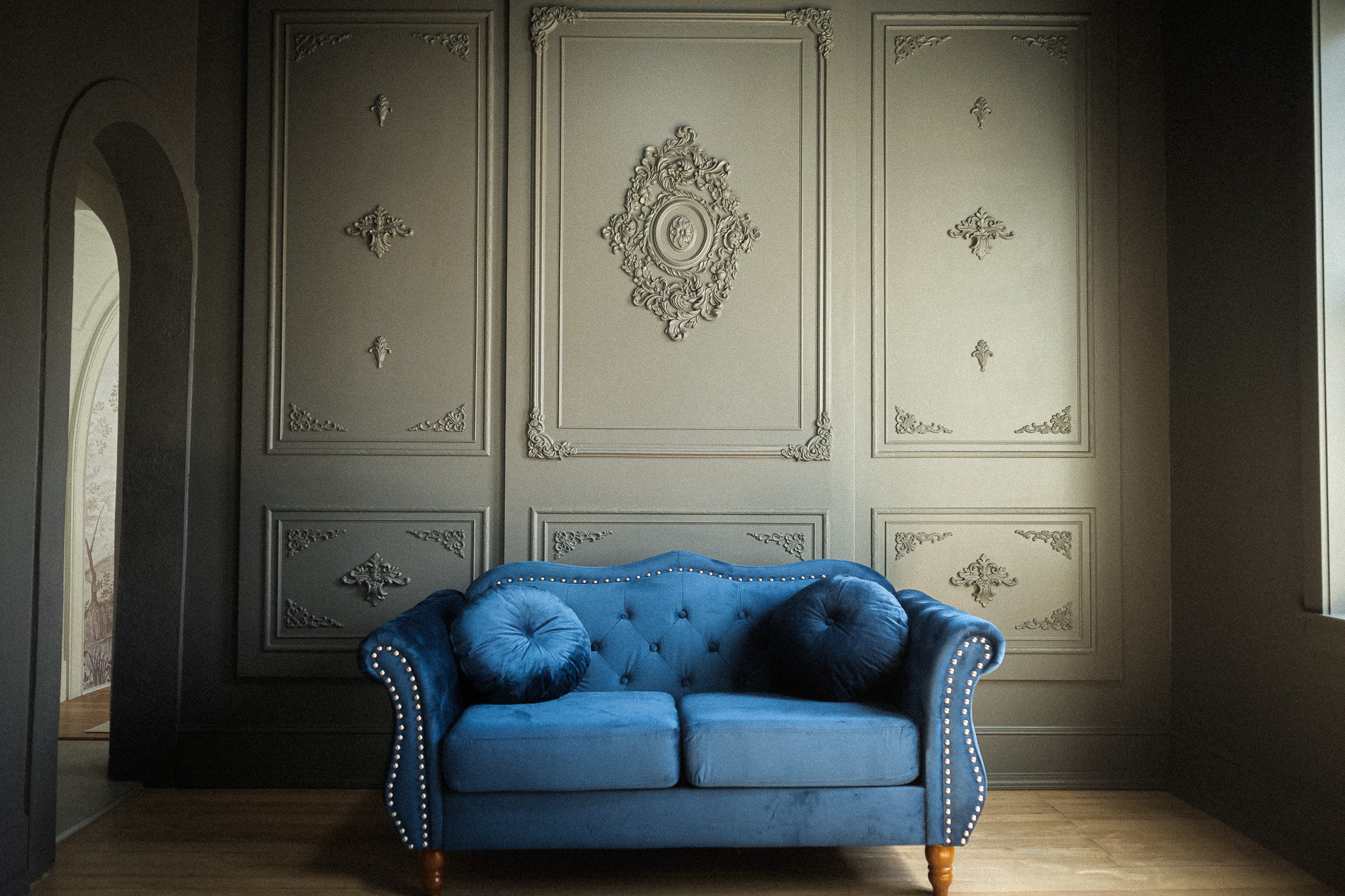 A blue tufted velvet sofa with round pillows on a wooden floor in front of an ornate, light gray wall with decorative molding.