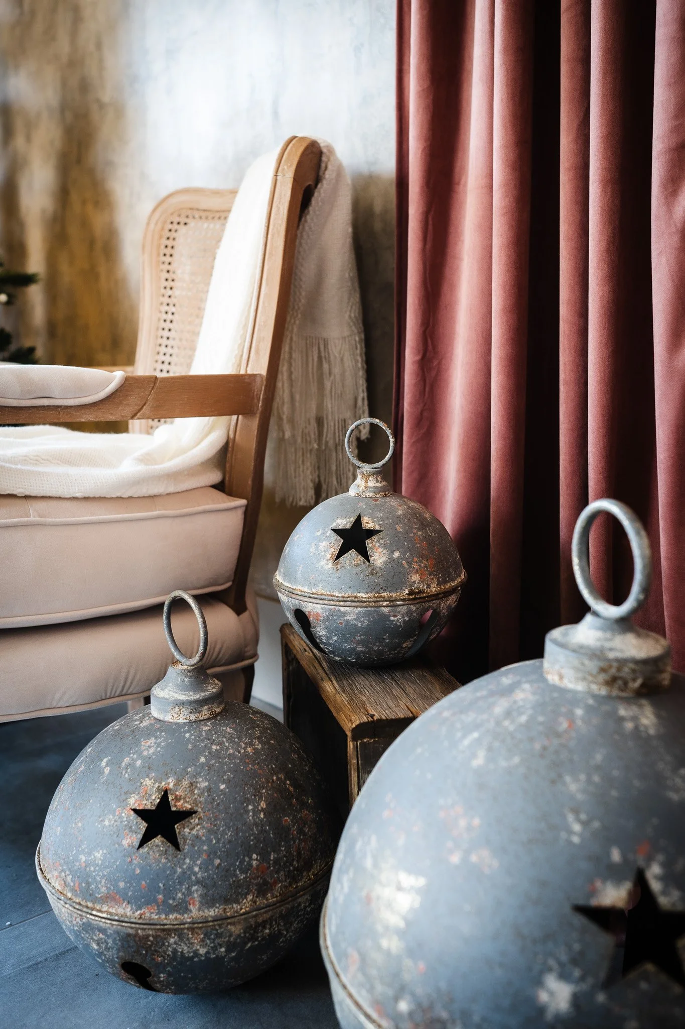 Vintage metal Christmas ornaments with star cutouts, rusted and distressed, placed on a wooden stand next to a cream-colored armchair and burgundy curtains.