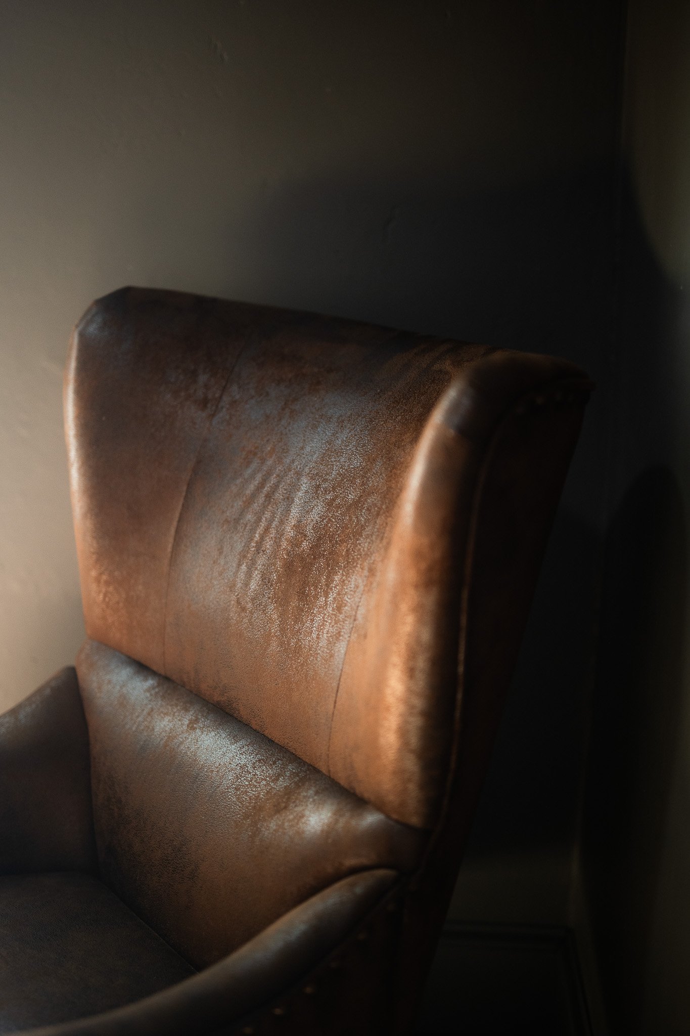 Close-up of an old, worn leather armchair with a high back, showing signs of age and extensive use, against a dark background.