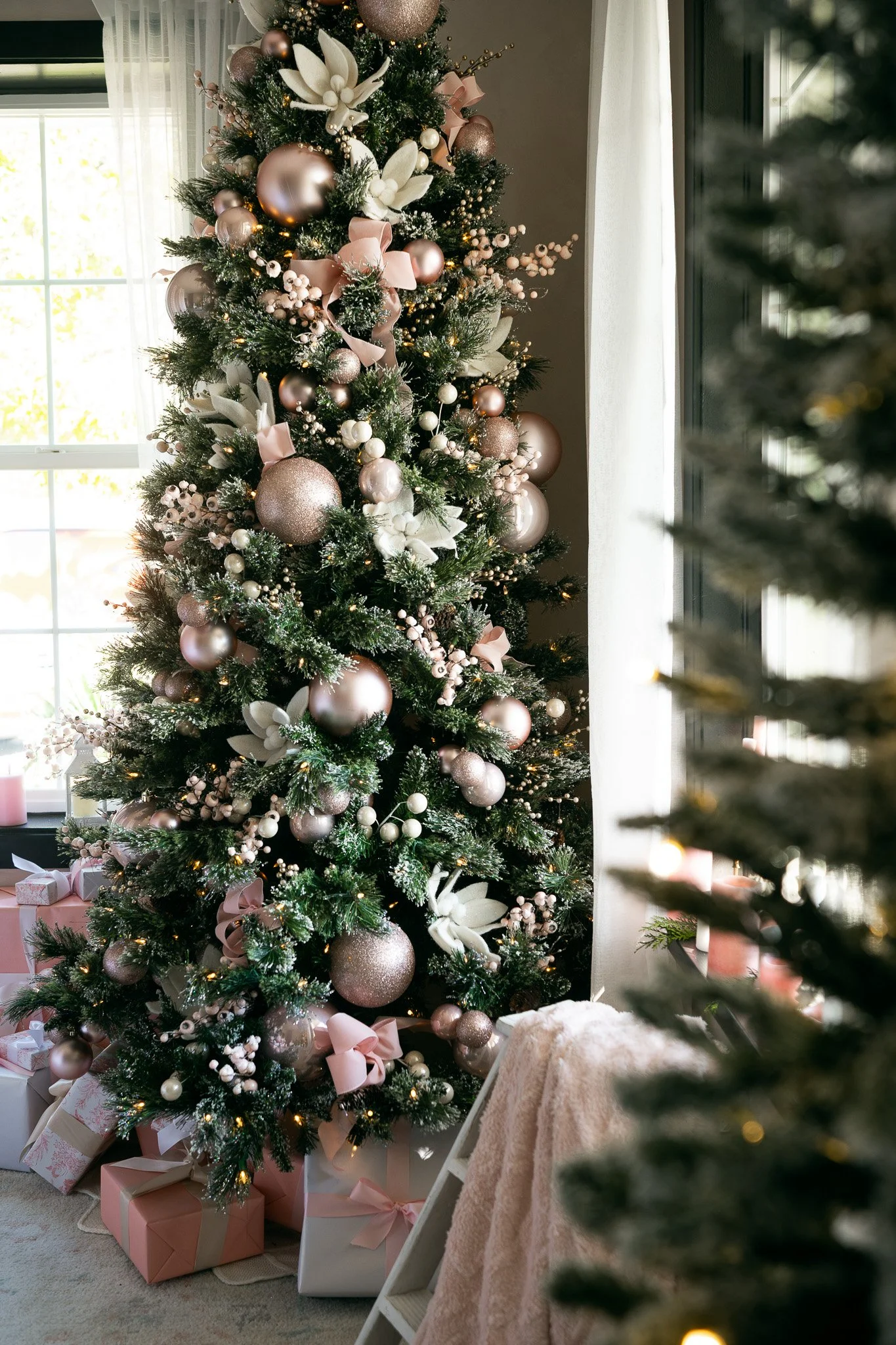 A decorated Christmas tree with pink, white, and gold ornaments, ribbons, and lights, with wrapped presents underneath, near a window with sheer curtains.