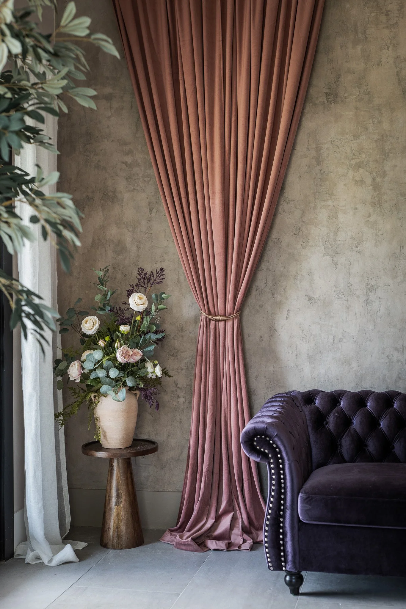 Interior corner with pink curtain, purple velvet sofa, and a flower arrangement in a beige vase on a wooden stand.
