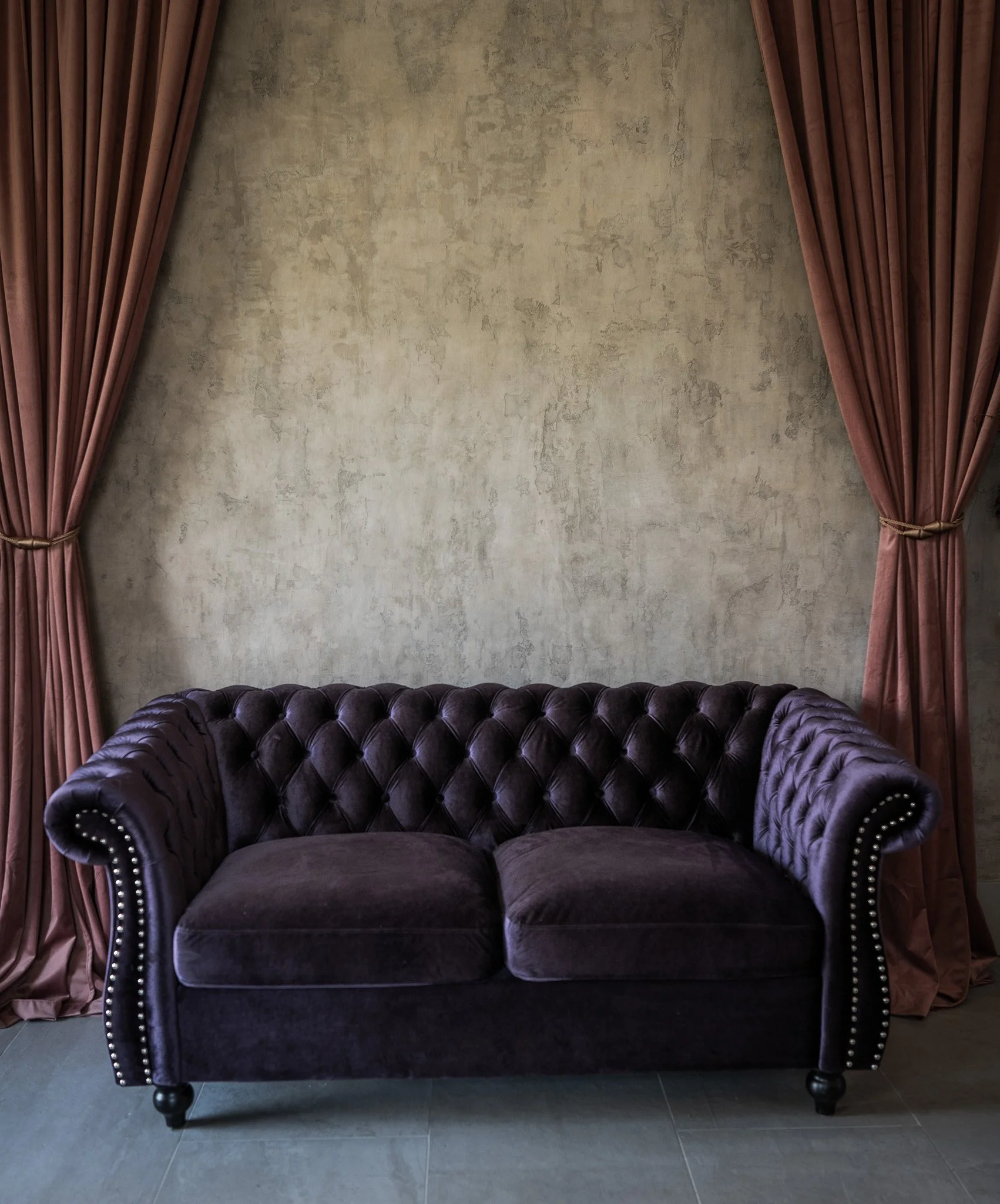 A dark purple velvet tufted sofa with nailhead trim on arms, set in front of an unfinished beige textured wall, with pinkish-brown curtains on either side.