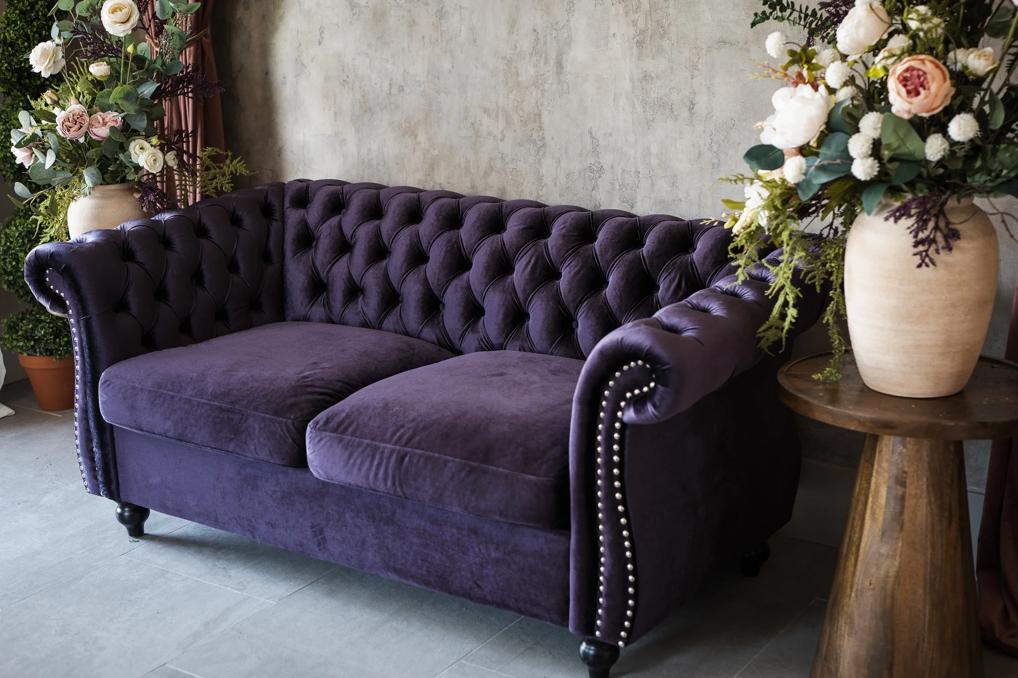 A dark purple velvet tufted sofa with nailhead trim, positioned against a textured gray wall, with two large beige vases filled with flowers on either side. One vase has mostly white and pink flowers, and the other has white and purple flowers, with